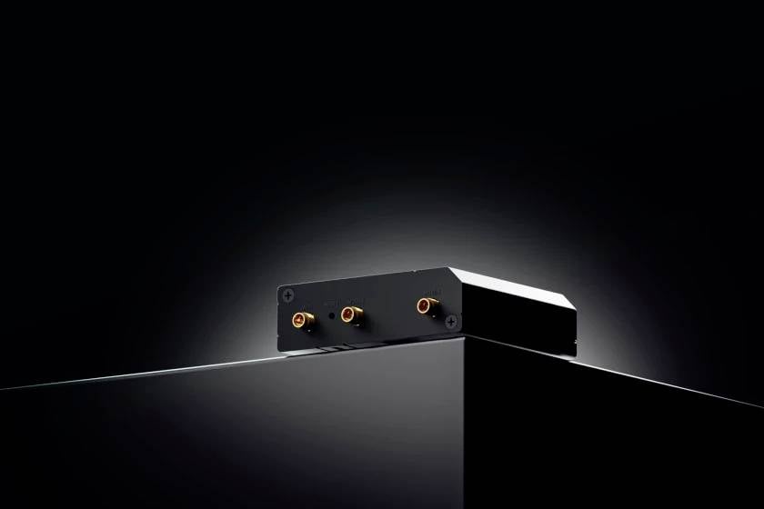 Ein schwarzes, rechteckiges elektronisches Gerät mit goldfarbenen Anschlüssen auf einer glänzenden schwarzen Oberfläche vor dunklem Hintergrund.