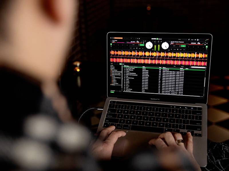 Eine Person arbeitet an einem Laptop, auf dem DJ-Software mit Musik-Visualisierung und Trackliste angezeigt wird.