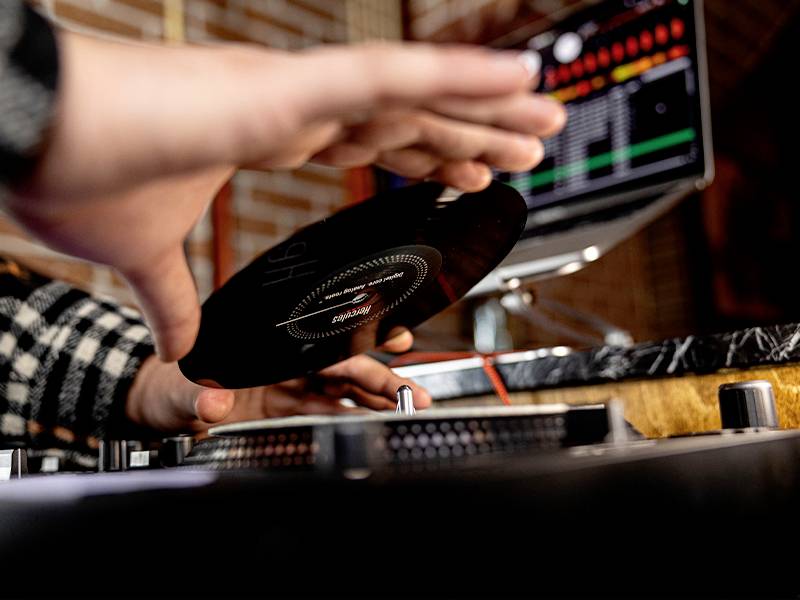 Eine Person legt eine Schallplatte auf einen Plattenspieler. Im Hintergrund ist ein Laptop mit Musik-Software sichtbar.