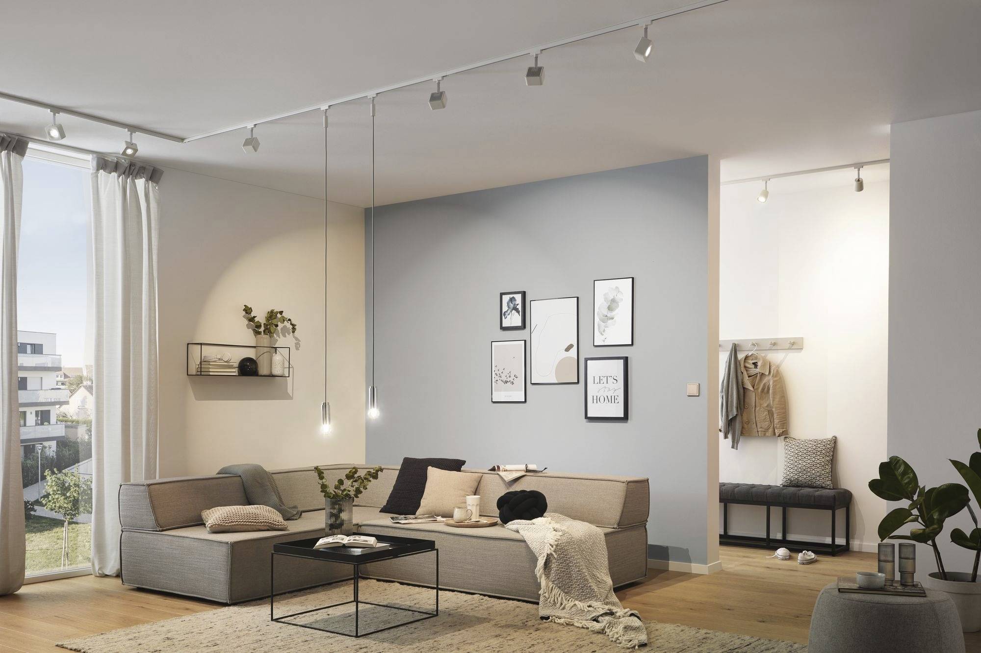 Ein modernes Wohnzimmer mit grauem Ecksofa, Couchtisch und hellblauer Akzentwand mit Bildern. Helles Tageslicht fällt durch ein großes Fenster.