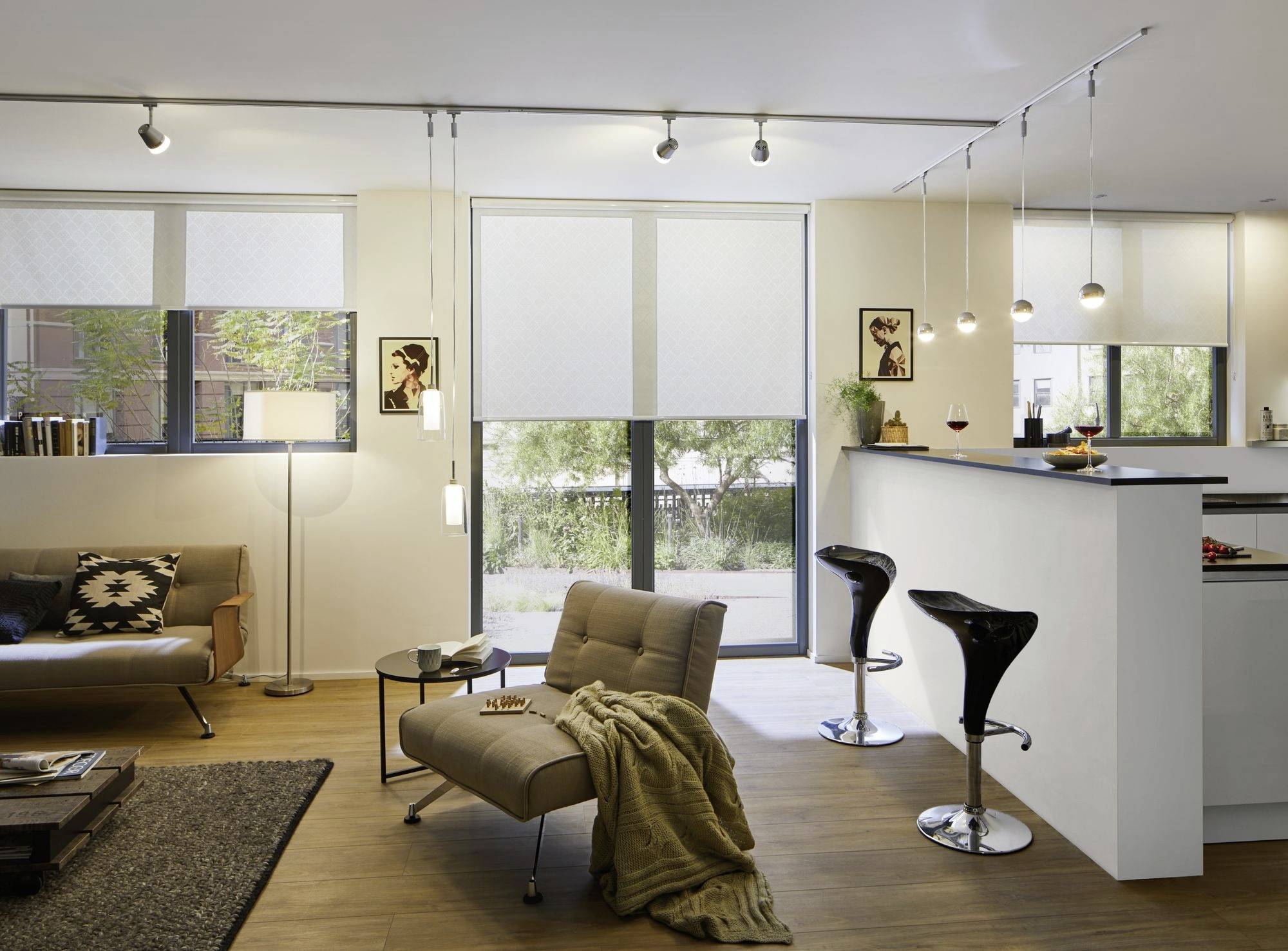 Modernes Wohnzimmer mit Holzfußboden, Sofa, Sessel, Deckenleuchten und Bar mit zwei Hockern; große Fenster im Hintergrund.