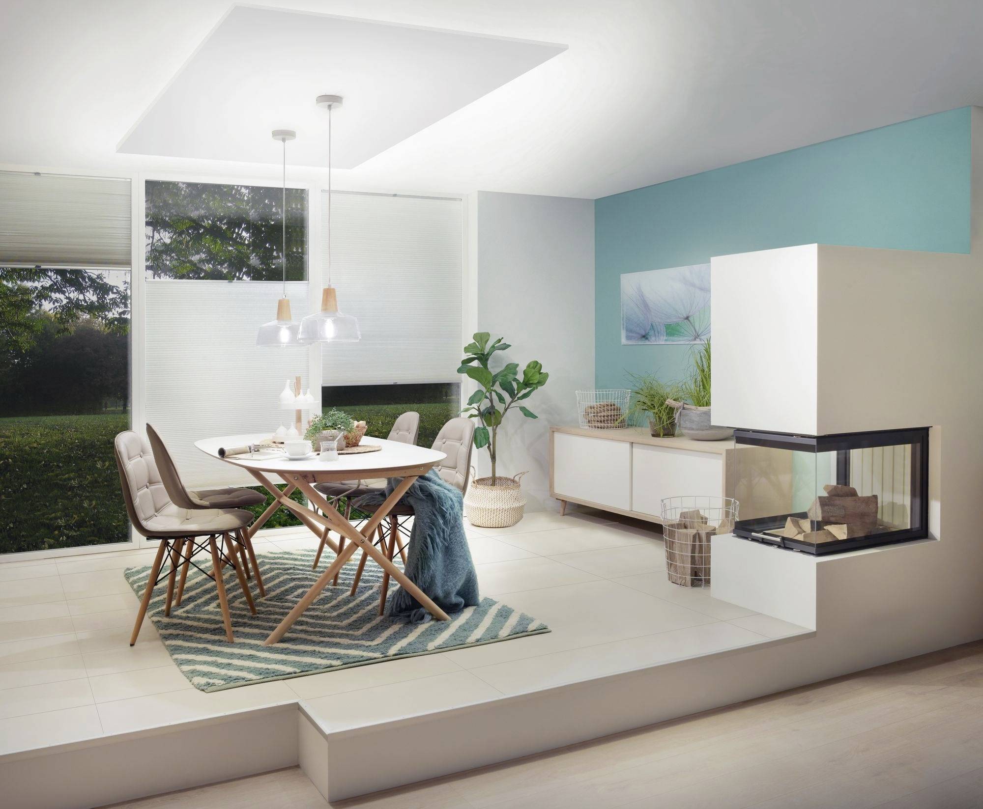 Esszimmer mit Esstisch und Stühlen, ein grünes Pflanzenarrangement neben einem modernen Kamin, große Fenster im Hintergrund.