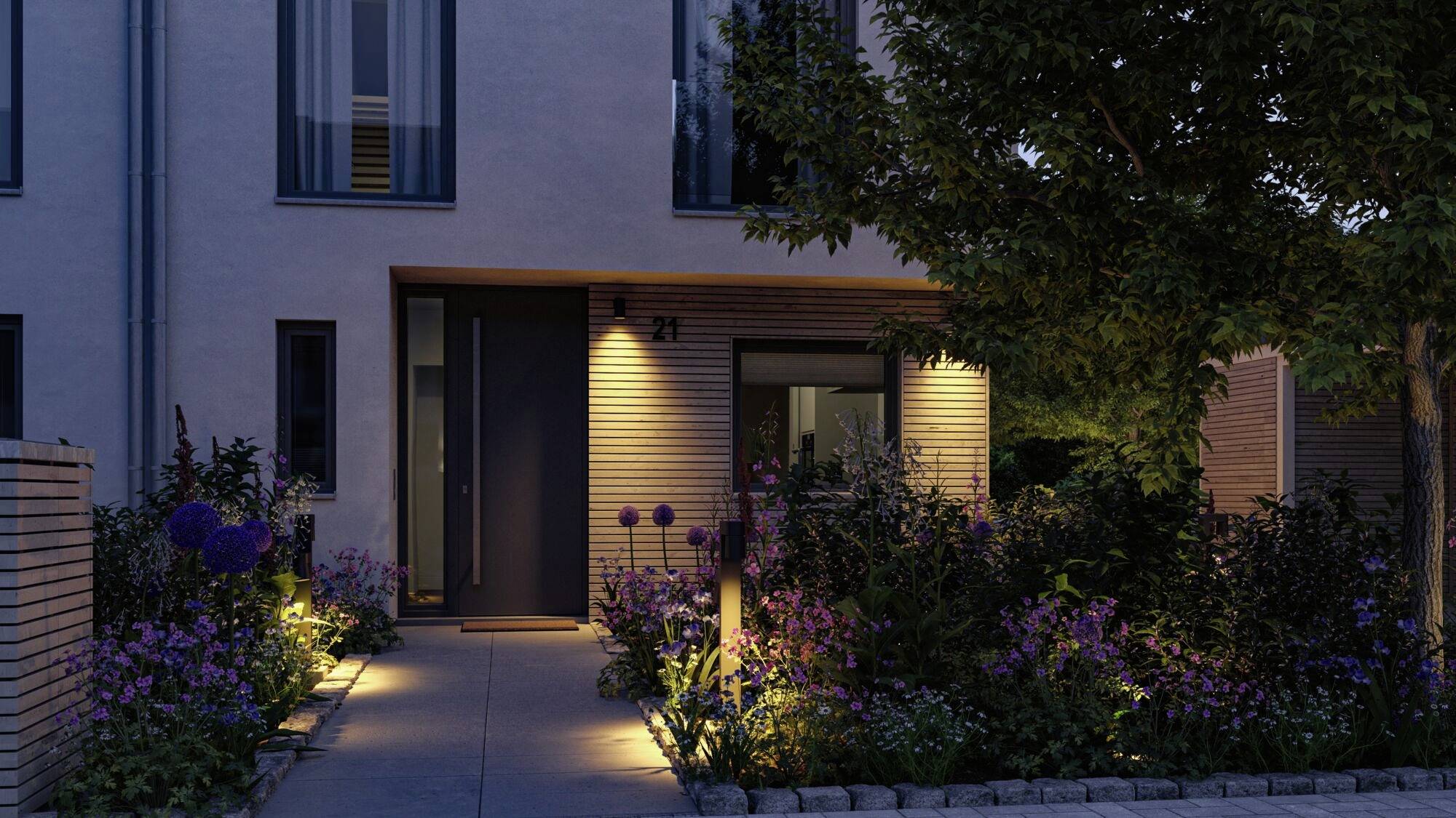 Eingangsbereich eines modernen Hauses am Abend, beleuchtet mit warmen Lichtern, umgeben von bunten Blumen und einem Baum rechts.