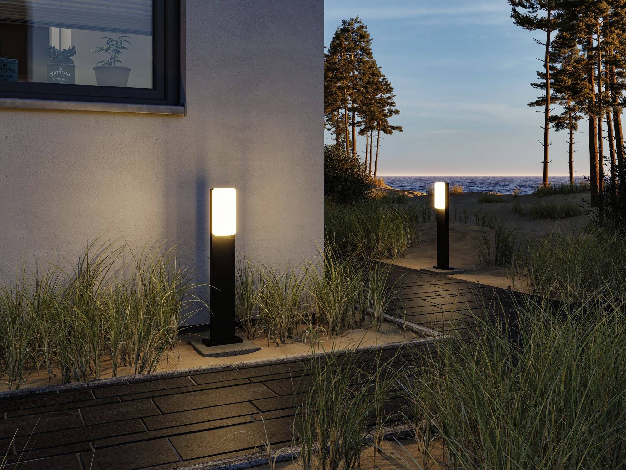 Ein beleuchteter Gartenweg führt an einem Gebäude entlang und endet in der Ferne am Meer. Gräser umgeben den Weg, der mit Lampen gesäumt ist.