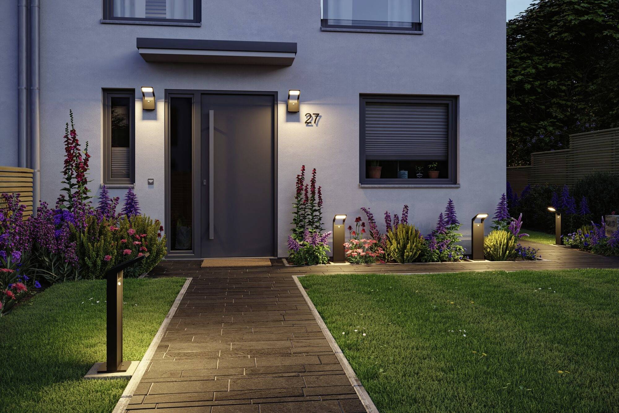 Ein modernes Haus mit beleuchtetem Eingang und gepflegtem Garten. Blumenbeete flankieren den Weg zur Haustür.