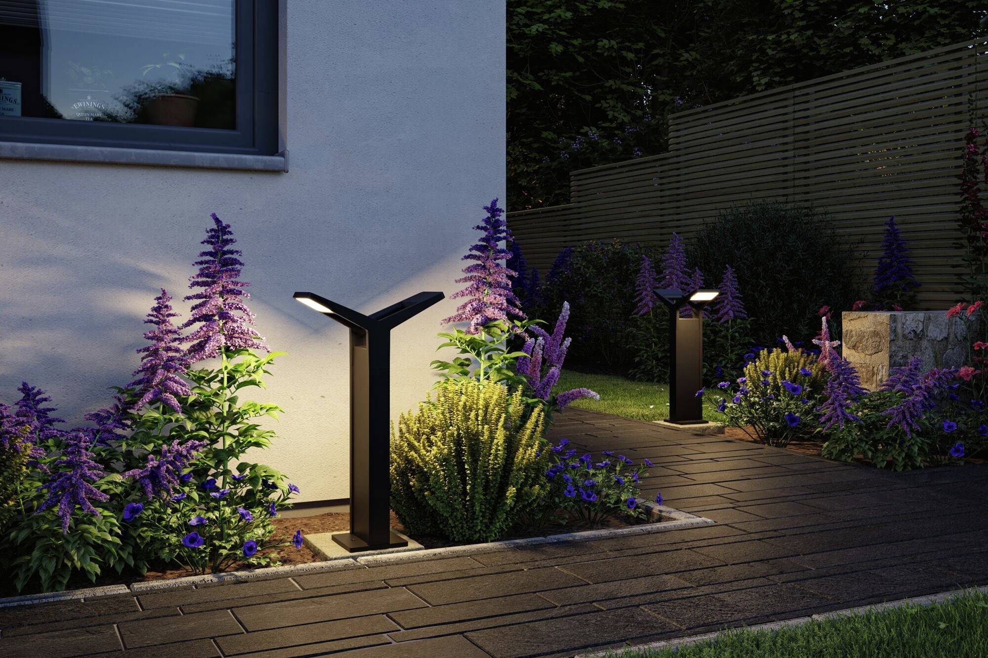 Gartenweg bei Nacht mit moderner Beleuchtung und Blumenbeeten. Kleiner gepflasterter Pfad führt entlang eines Hauses, umgeben von lila Blumen.