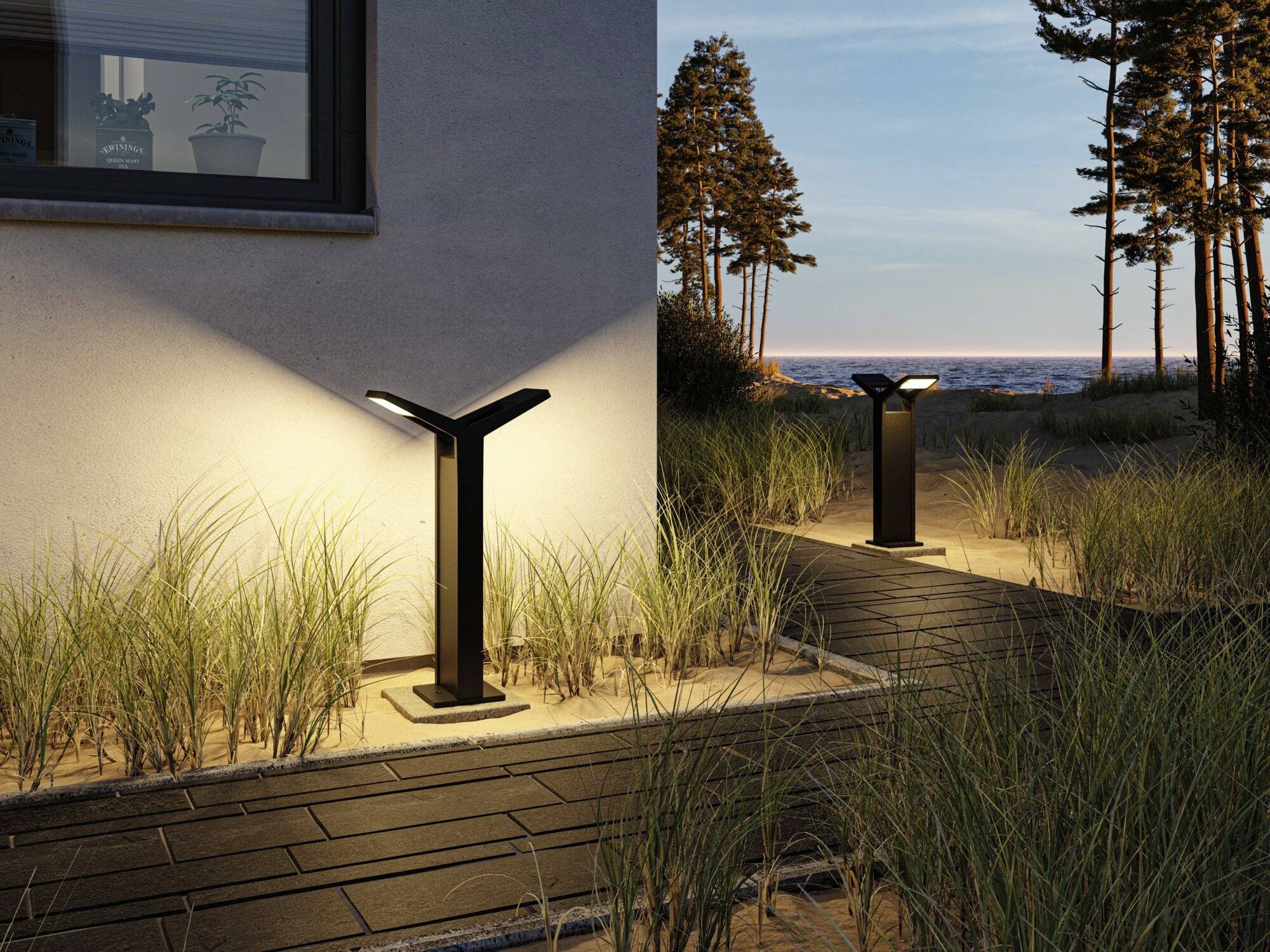 Moderne Außenleuchten beleuchten einen Gehweg neben einem Haus. Im Hintergrund ist ein bewaldetes Gebiet und das Meer sichtbar.