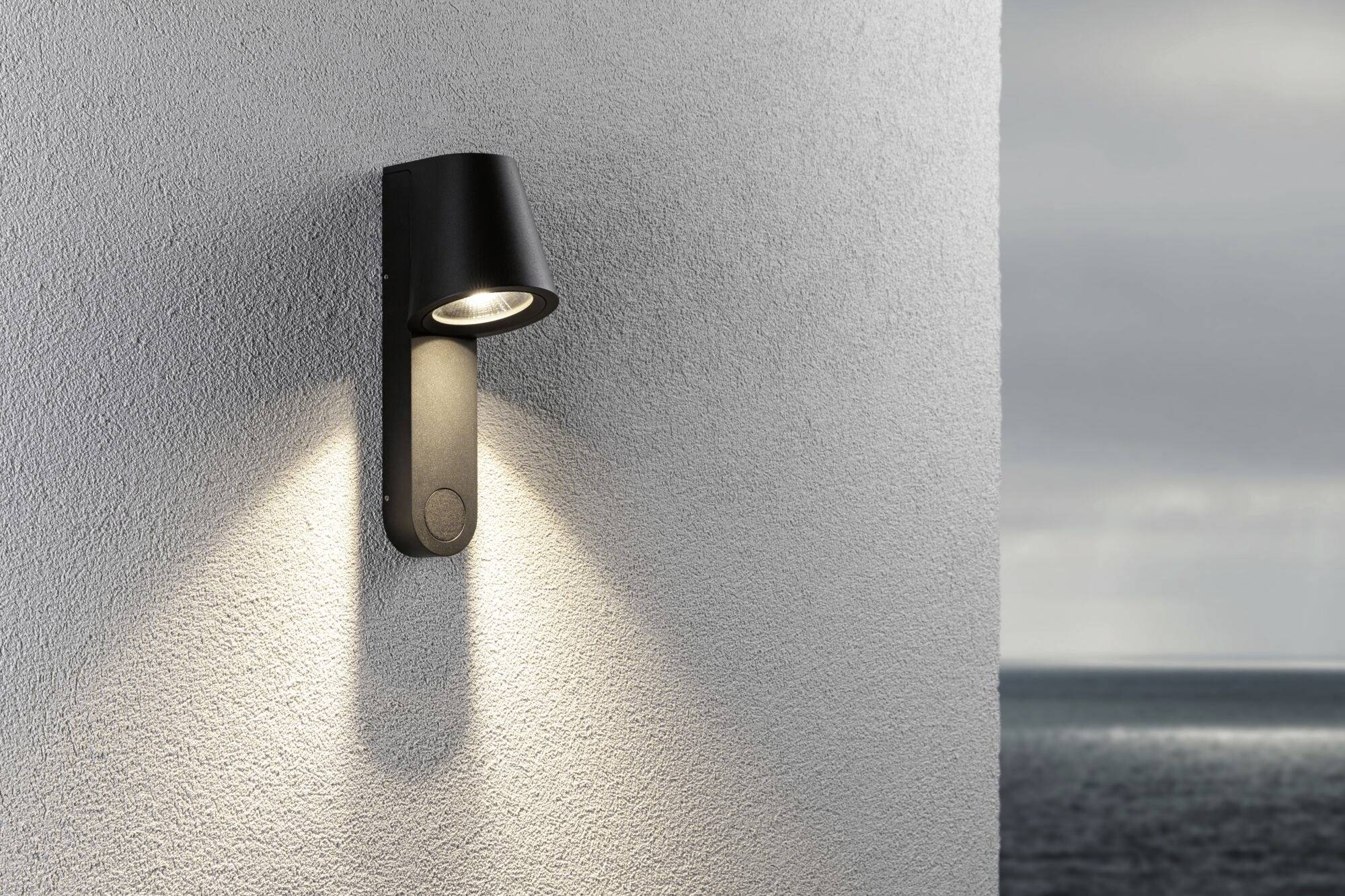Außenleuchte an einer Hauswand, strahlt warmes Licht nach unten. Im Hintergrund ist ein wolkiger Himmel über dem Meer zu sehen.