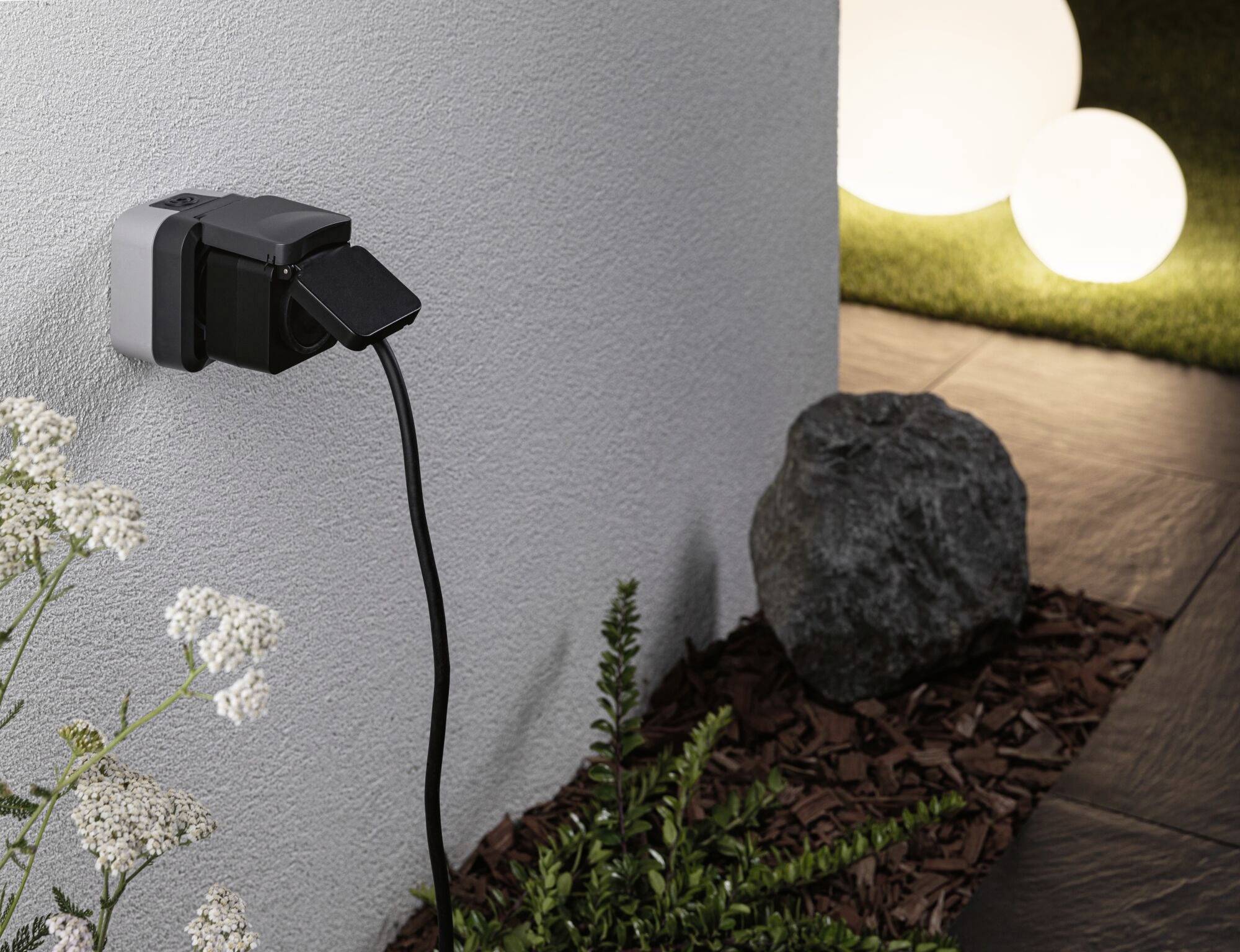 Steckdose an Außenwand mit eingestecktem schwarzen Stecker; Pflanzen und Steine im Vordergrund, leuchtende Kugeln im Hintergrund.