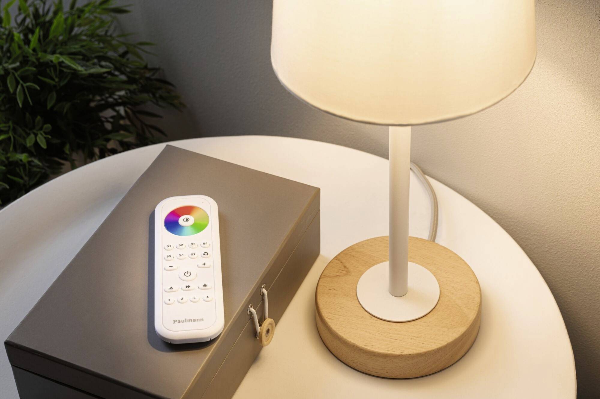 Eine Tischlampe mit Holzsockel leuchtet auf einem runden Tisch. Daneben liegt eine Fernbedienung mit Farbkreis, vor einer grauen Box.