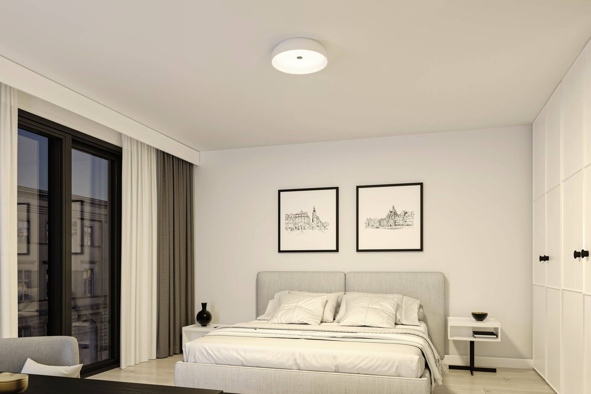 Modernes Schlafzimmer mit einem grauen Doppelbett, zwei gerahmten Kunstwerken an der Wand, einem kleinen Nachttisch und einem großen Fenster.