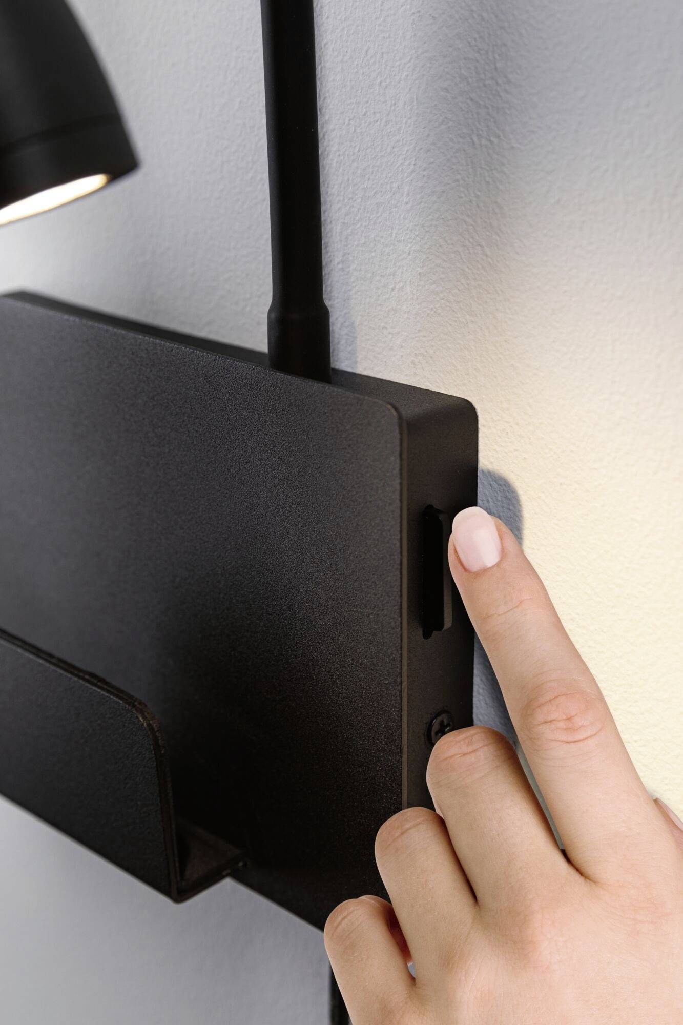 Eine Hand berührt eine schwarze Box an der Wand, möglicherweise eine Schaltfläche oder einen Schalter. Eine Lampe ist in Nähe sichtbar.