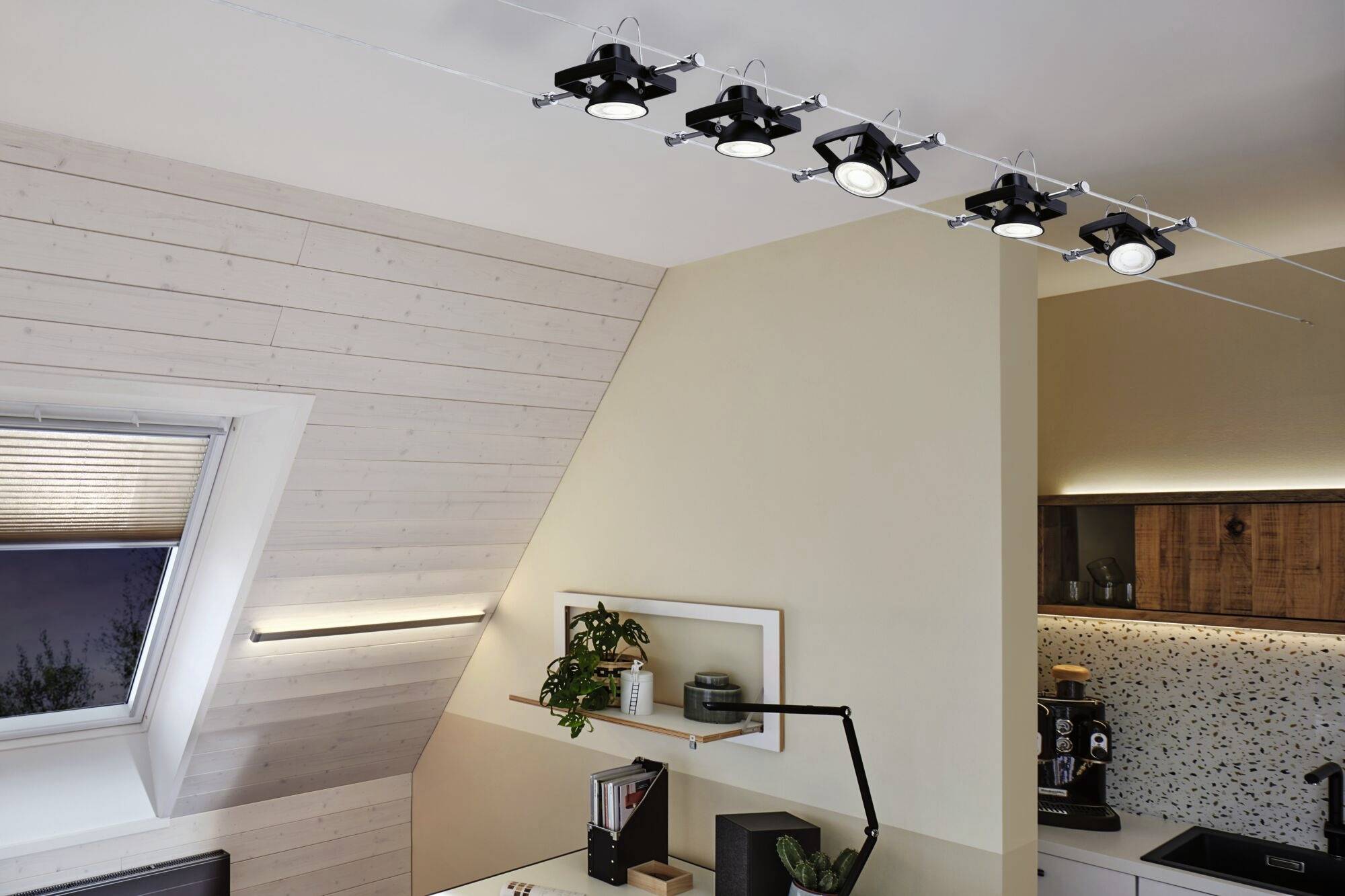 Ein modernes Wohnzimmer mit hellem Holzdekor und schrägen Wänden. Eine Reihe von neun schwarzen Deckenstrahlern beleuchtet den Raum.