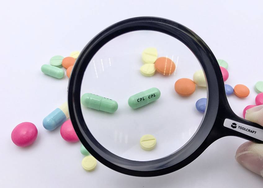 Verschiedene bunte Tabletten, einige vergrößert durch eine Lupe. Beige Hintergrund, Brennpunkt auf zwei grüne Kapseln mit Aufschrift 'CPL'.