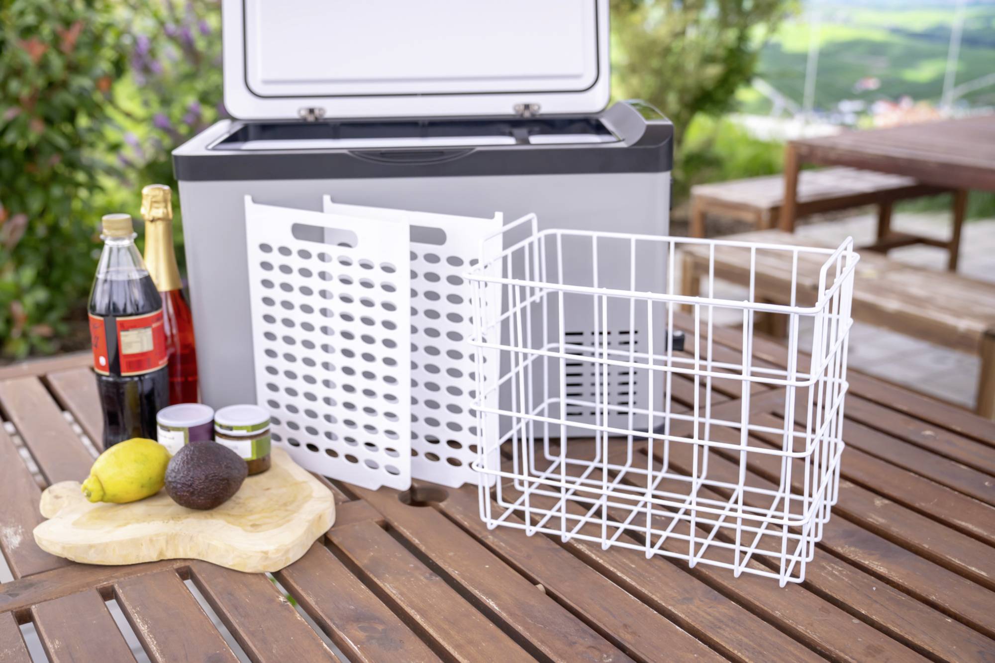 Kühlbox auf Holztisch im Freien mit Cola, Sekt, Avocado, Zitrone, Marmelade und Drahtkorb. Hintergrund zeigt verschwommene Pflanzen.