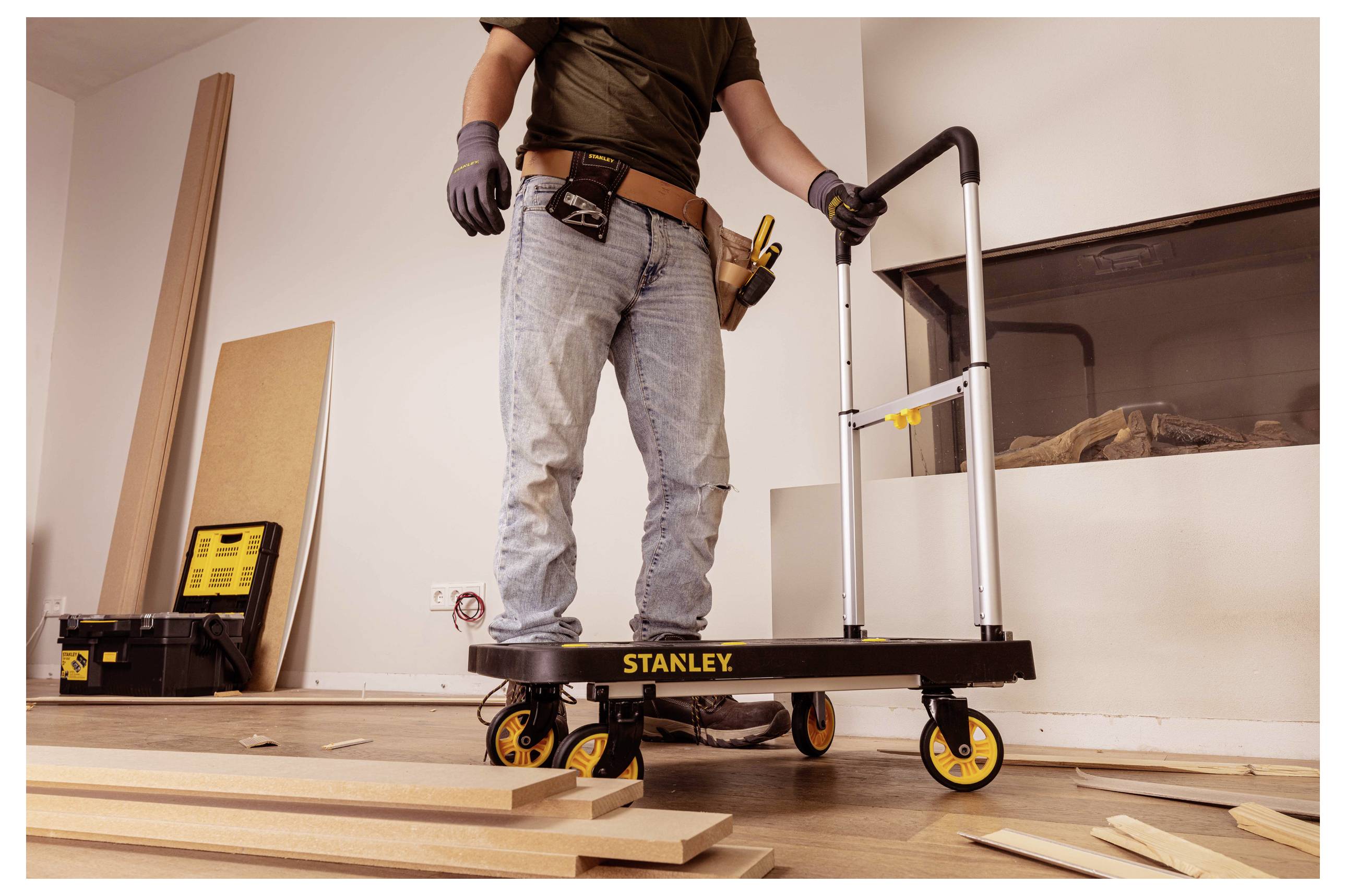 Eine Person in Arbeitskleidung steht auf einer fahrbaren Plattform in einer Baustelle, umgeben von Holzbrettern und Werkzeugen.
