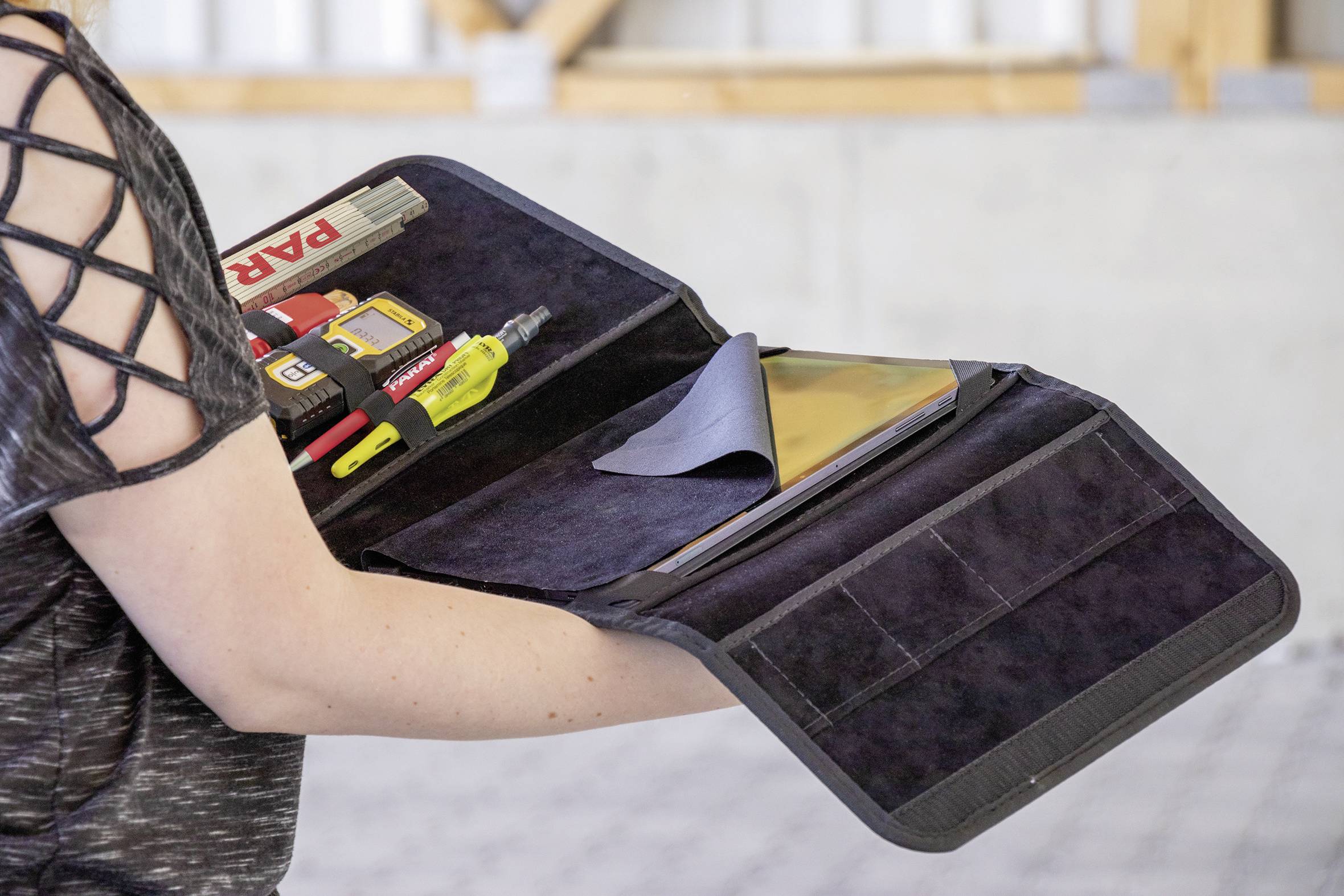 Eine Person hält eine offene Werkzeugtasche. Innen sind ein Maßband, ein Cutter und weitere Werkzeuge sichtbar.