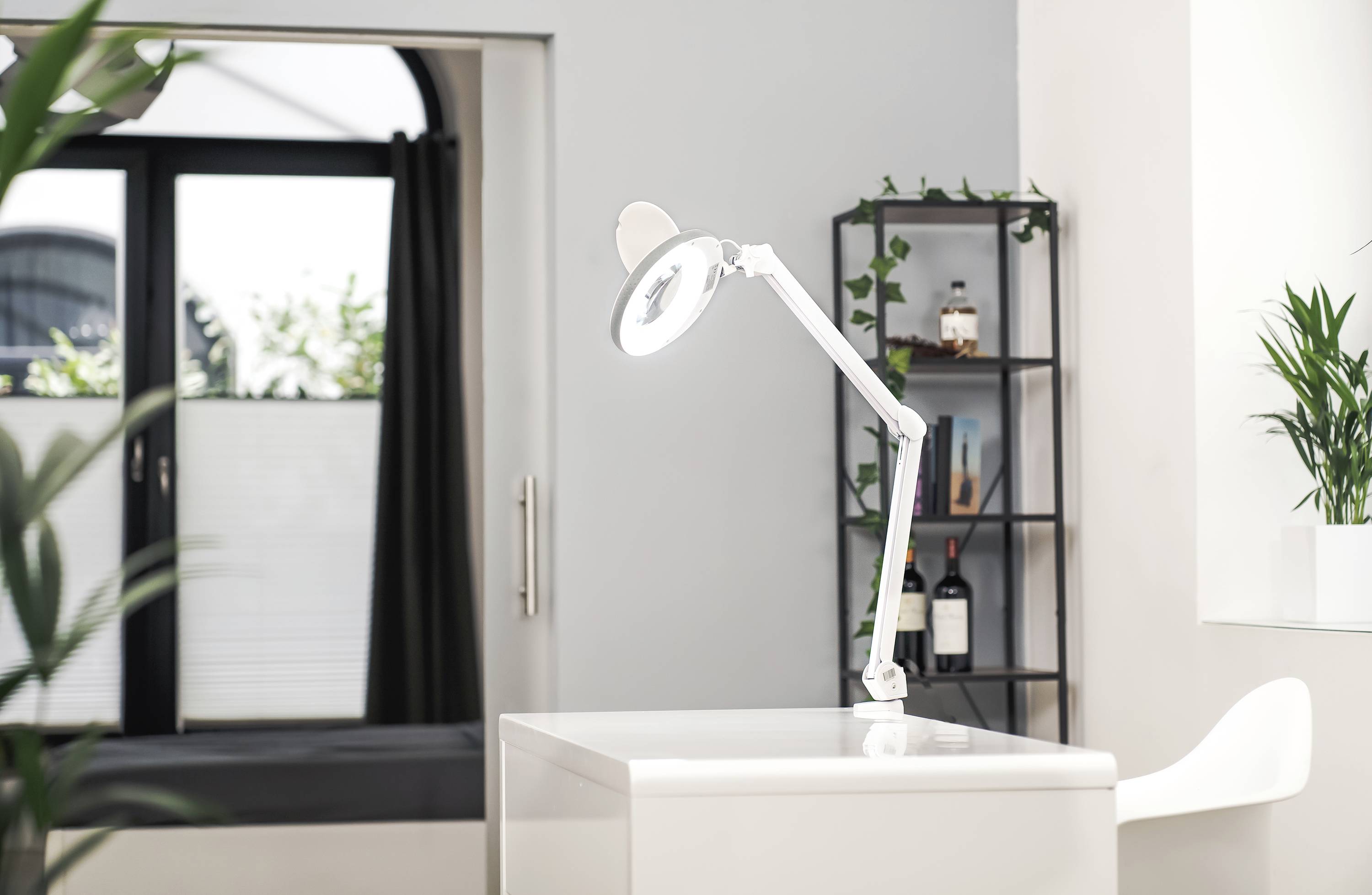 Ein minimalistischer Schreibtisch mit weißer Lampe; im Hintergrund ein Regal mit Pflanzen und Flaschen; helle Raumatmosphäre.