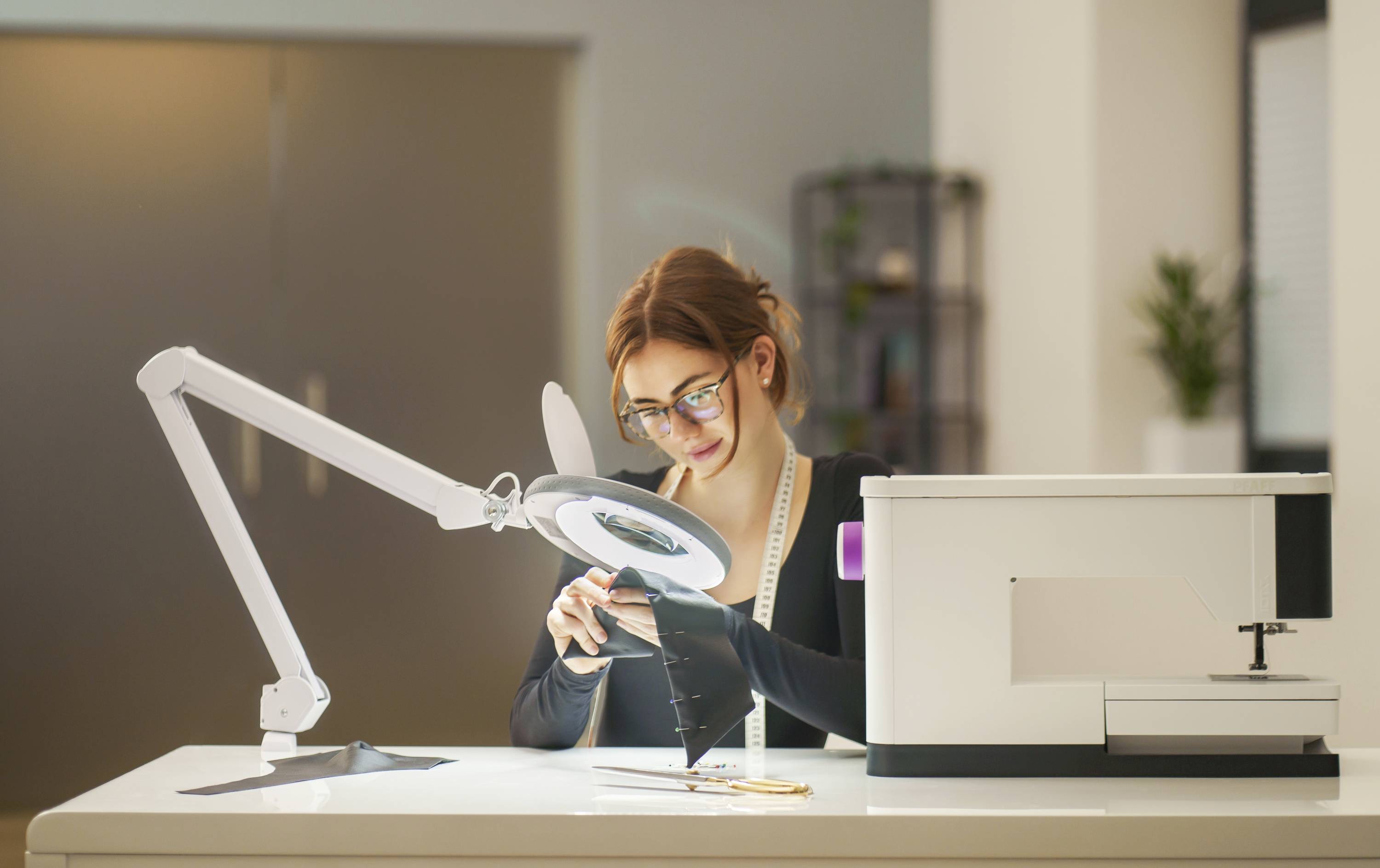 Eine Frau arbeitet konzentriert an einem technischen Gerät an einem Tisch in einem modernen Büro. Sie scheint ein Elektronikteil zu inspizieren.