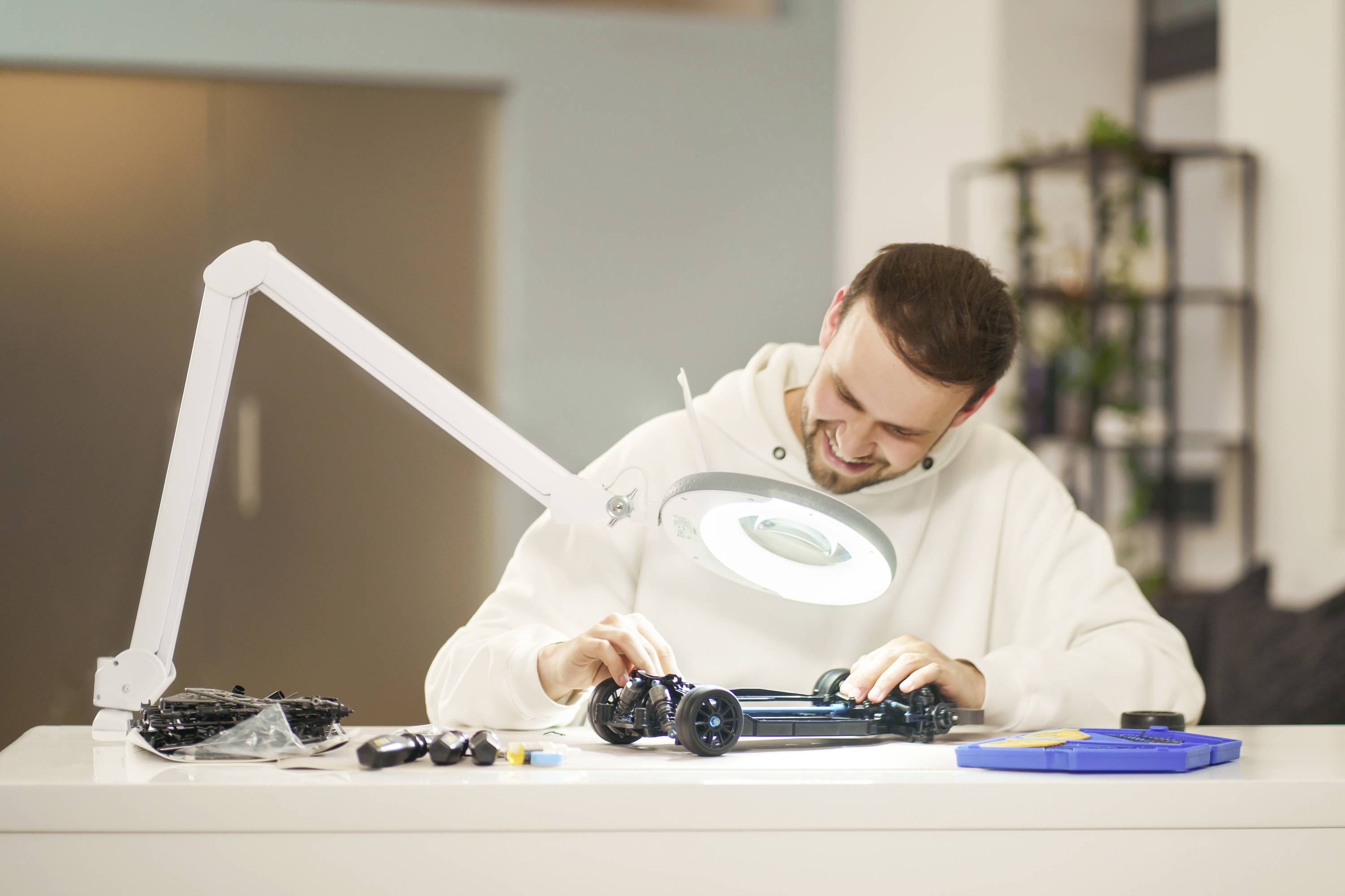 Ein Mann baut ein Modellauto an einem Tisch zusammen, beleuchtet von einer großen, runden Lampe. Um ihn sind Werkzeuge und Teile verstreut.