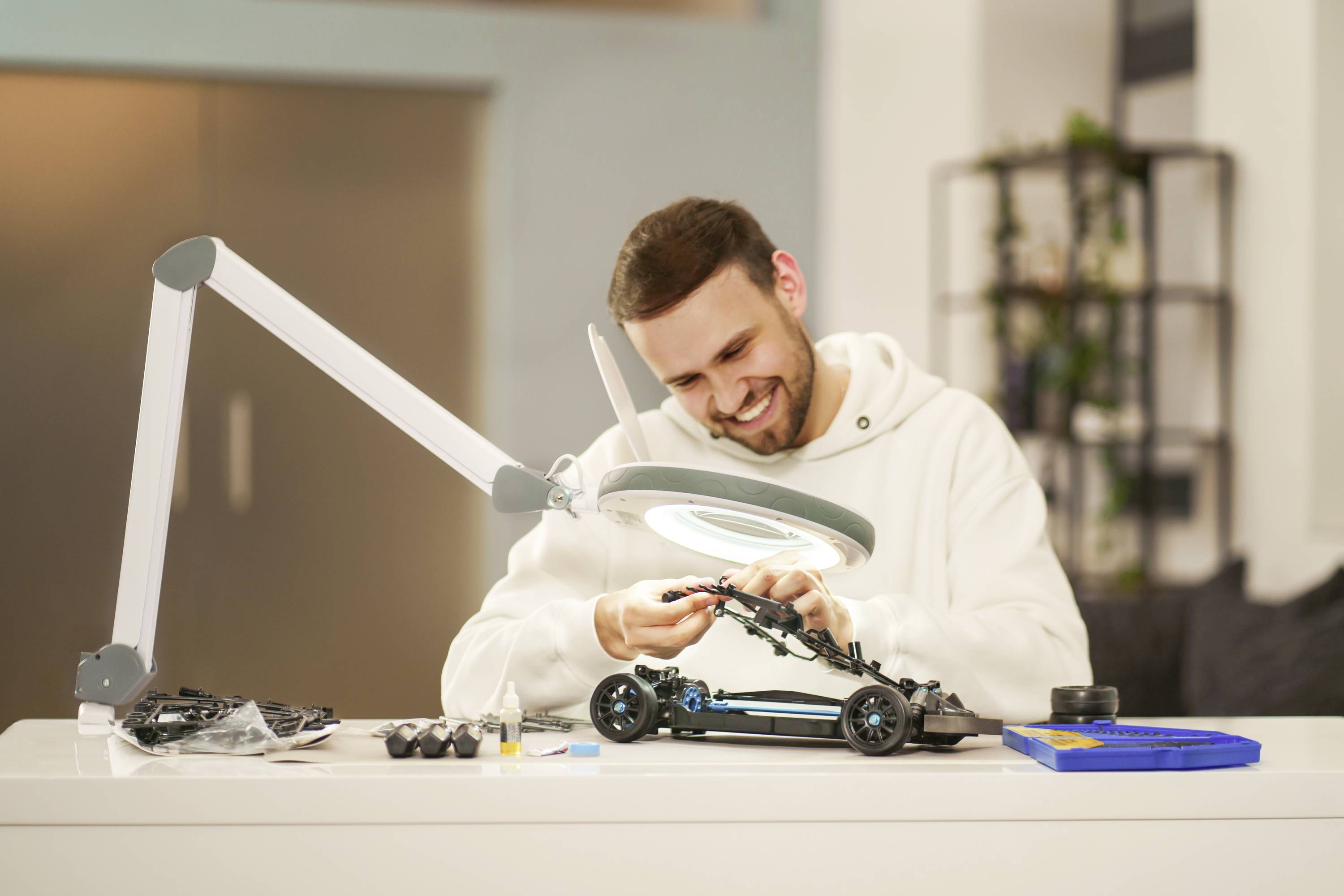 Ein Mann baut ein Modellauto an einem Tisch zusammen, umgeben von Werkzeugen und Teilen; er wirkt konzentriert und zufrieden.