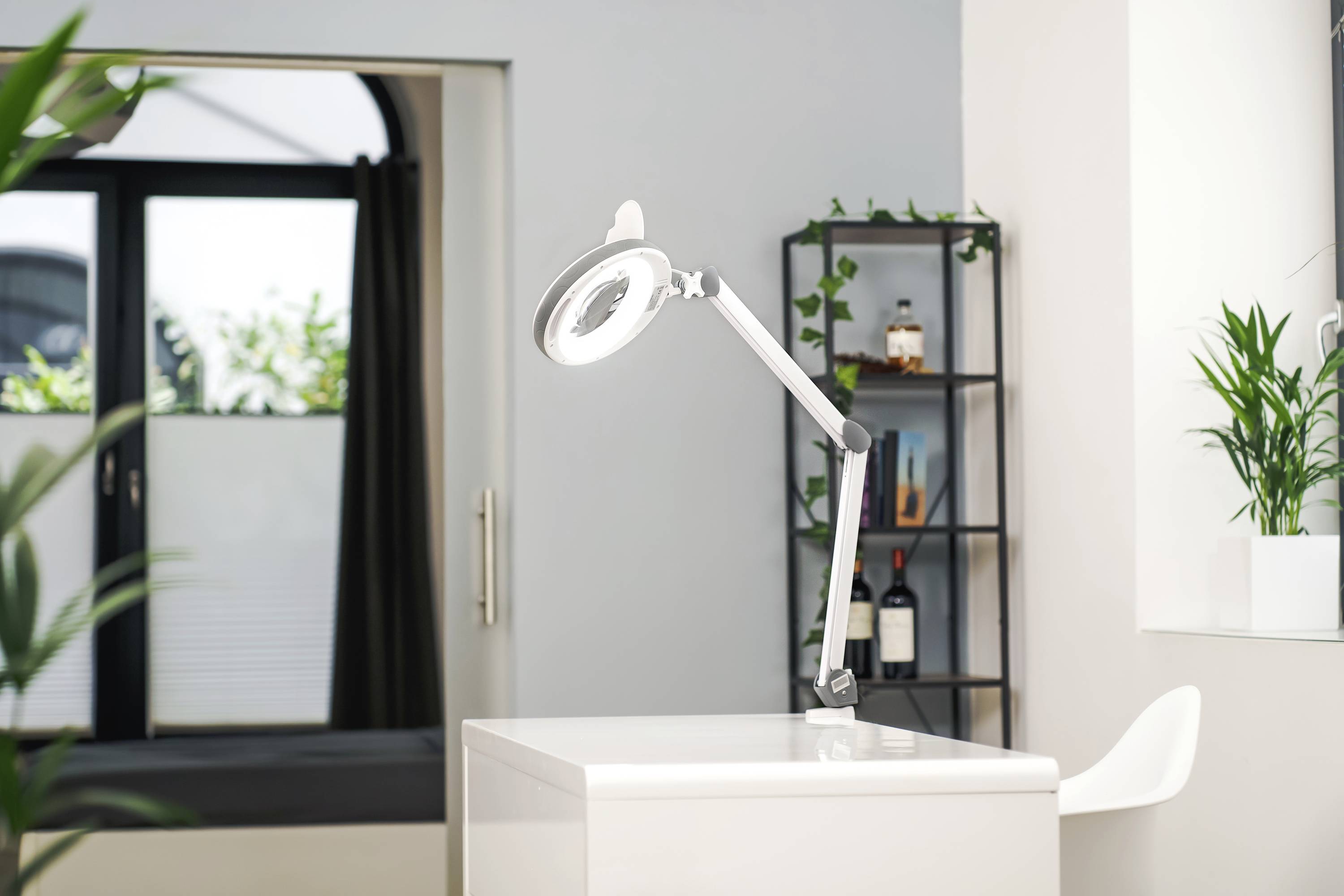 Helle Lampe mit Lupe auf einem weißen Tisch in modernem Raum. Hintergrund mit Pflanzen und Regal. Helle, ruhige Atmosphäre.