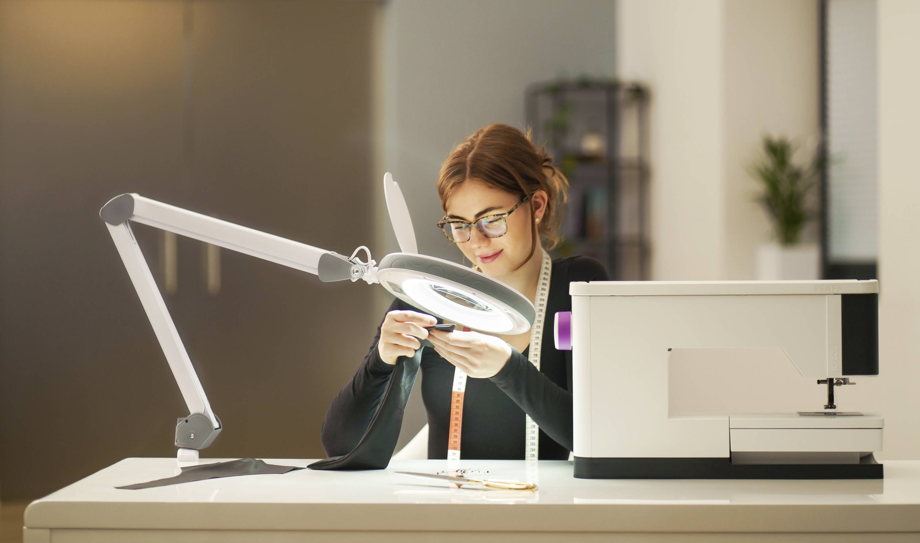 Eine Frau arbeitet konzentriert an einem Nähtisch mit einer Nähmaschine und einer Lupenlampe. Im Hintergrund sind Regale sichtbar.