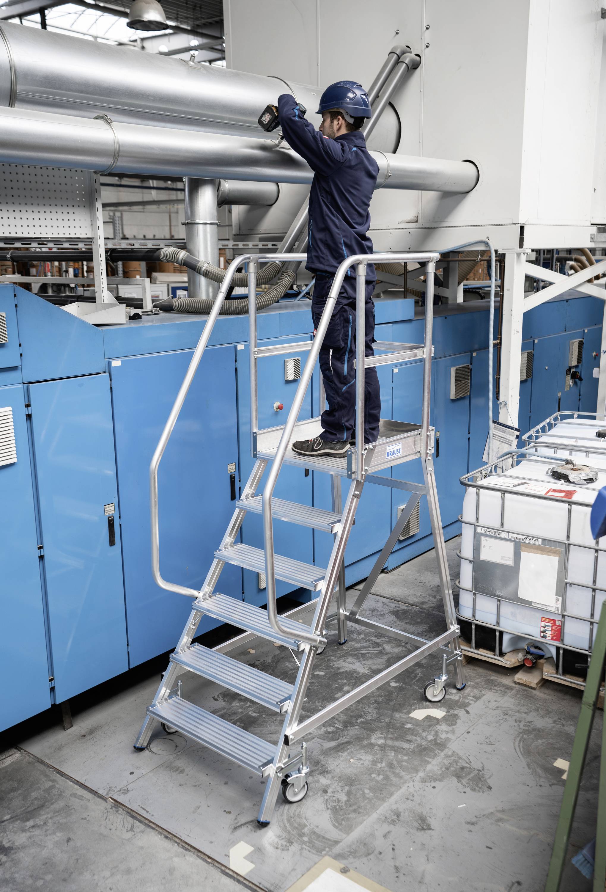 Ein Techniker in Sicherheitskleidung steht auf einer mobilen Treppe und wartet eine industrielle Lüftungsanlage in einer Fabrikumgebung.
