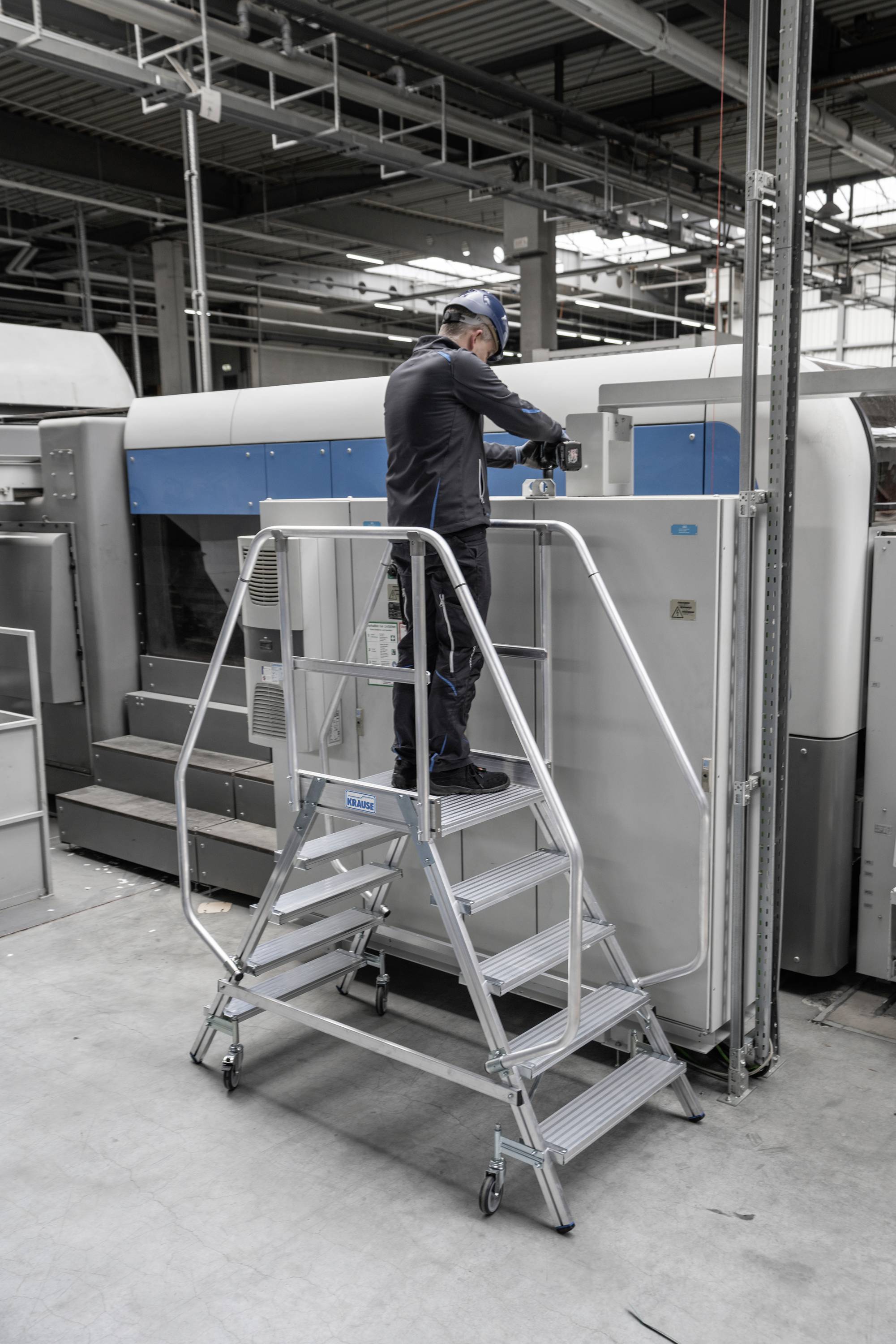 Ein Techniker arbeitet auf einer Metallplattform vor einem großen, industriellen Drucker in einer Fabrikumgebung.