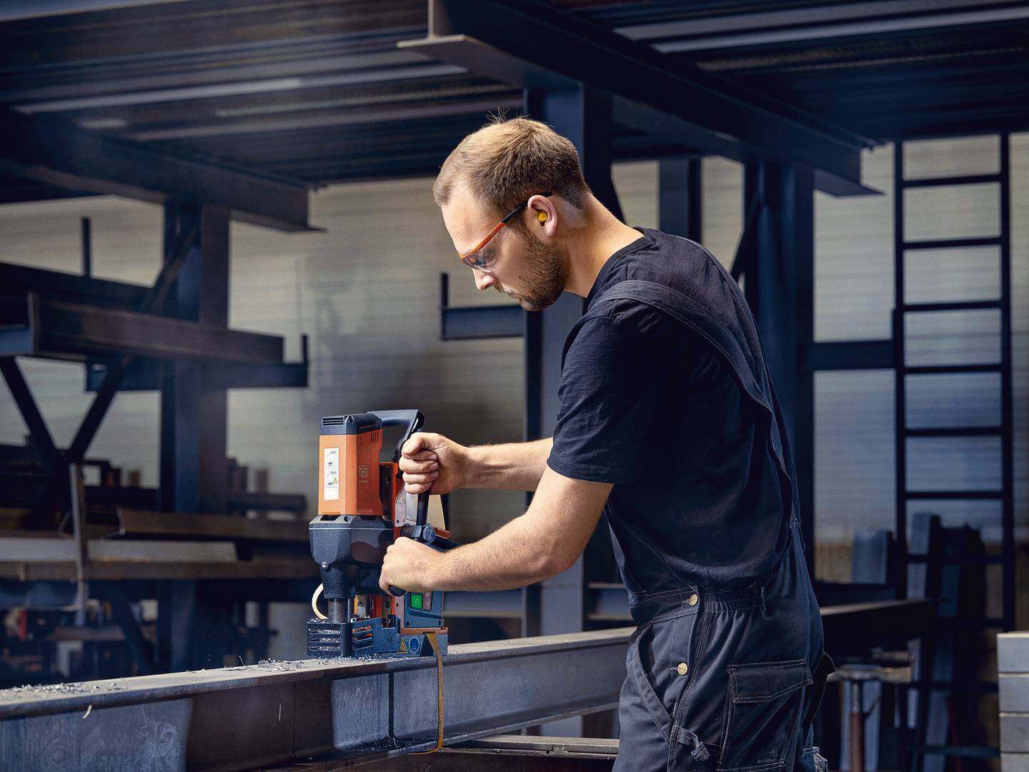 Ein Mann in Arbeitskleidung und Schutzbrille bohrt mit einer Maschine in einen Metallträger in einer Werkstatt oder Fabrikhalle.