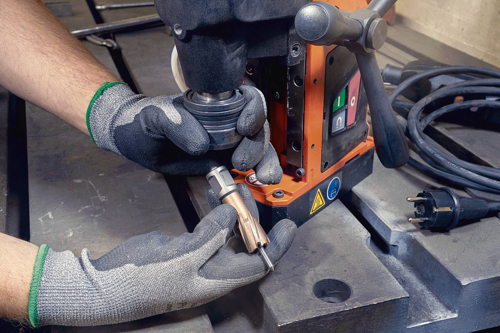 Eine Person mit Handschuhen bedient eine industrielle Bohrmaschine. Eine Bohrerspitze wird neben der Maschine gehalten.