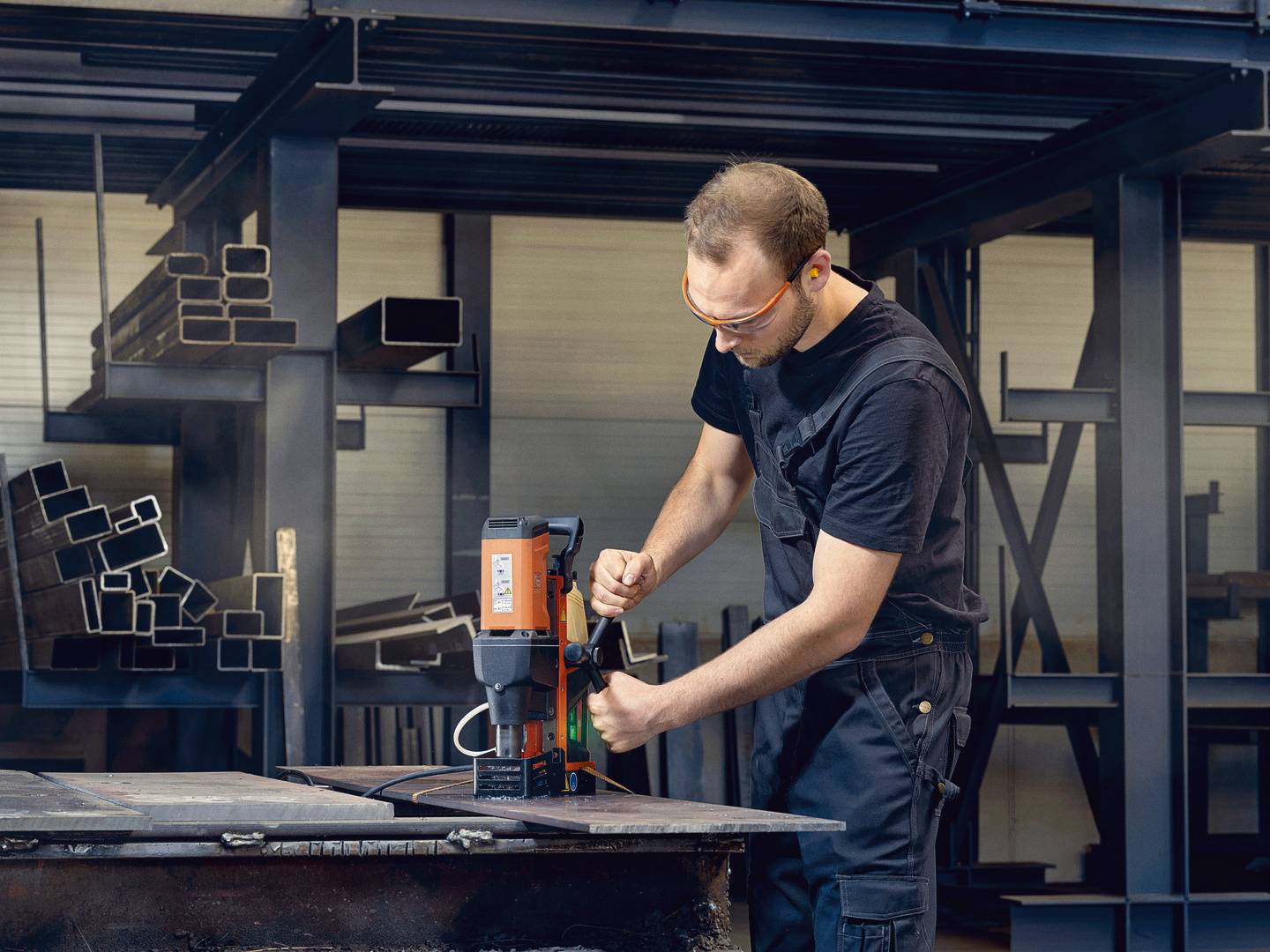Ein Mann in Schutzkleidung verwendet eine Bohrmaschine in einer Werkstatt, um Metallprofile auf einem Arbeitsbereich zu bearbeiten.