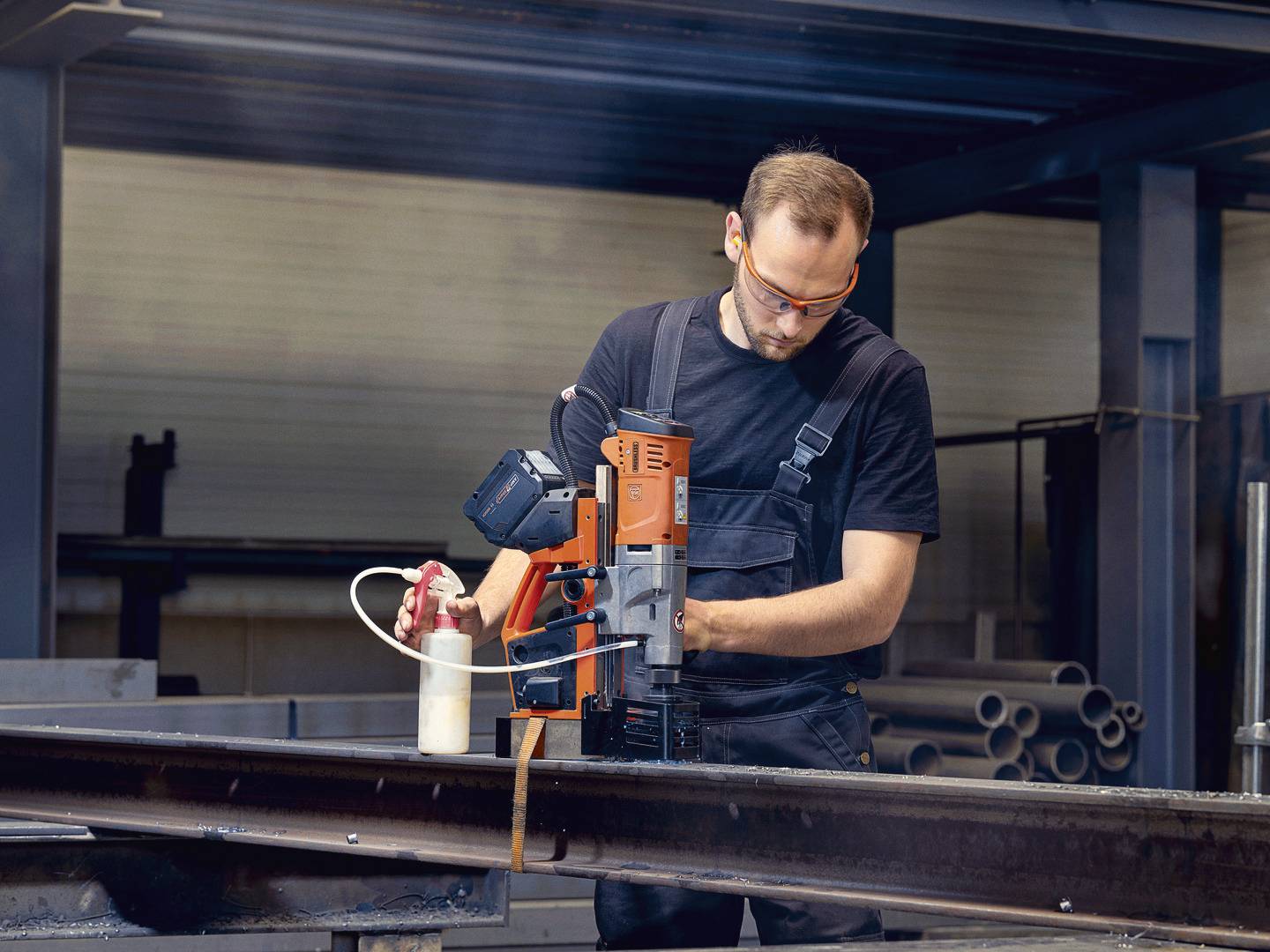 Mann in Arbeitskleidung benutzt eine Bohrmaschine auf einem Metallträger in einer Werkstattumgebung. Er trägt Schutzbrille.