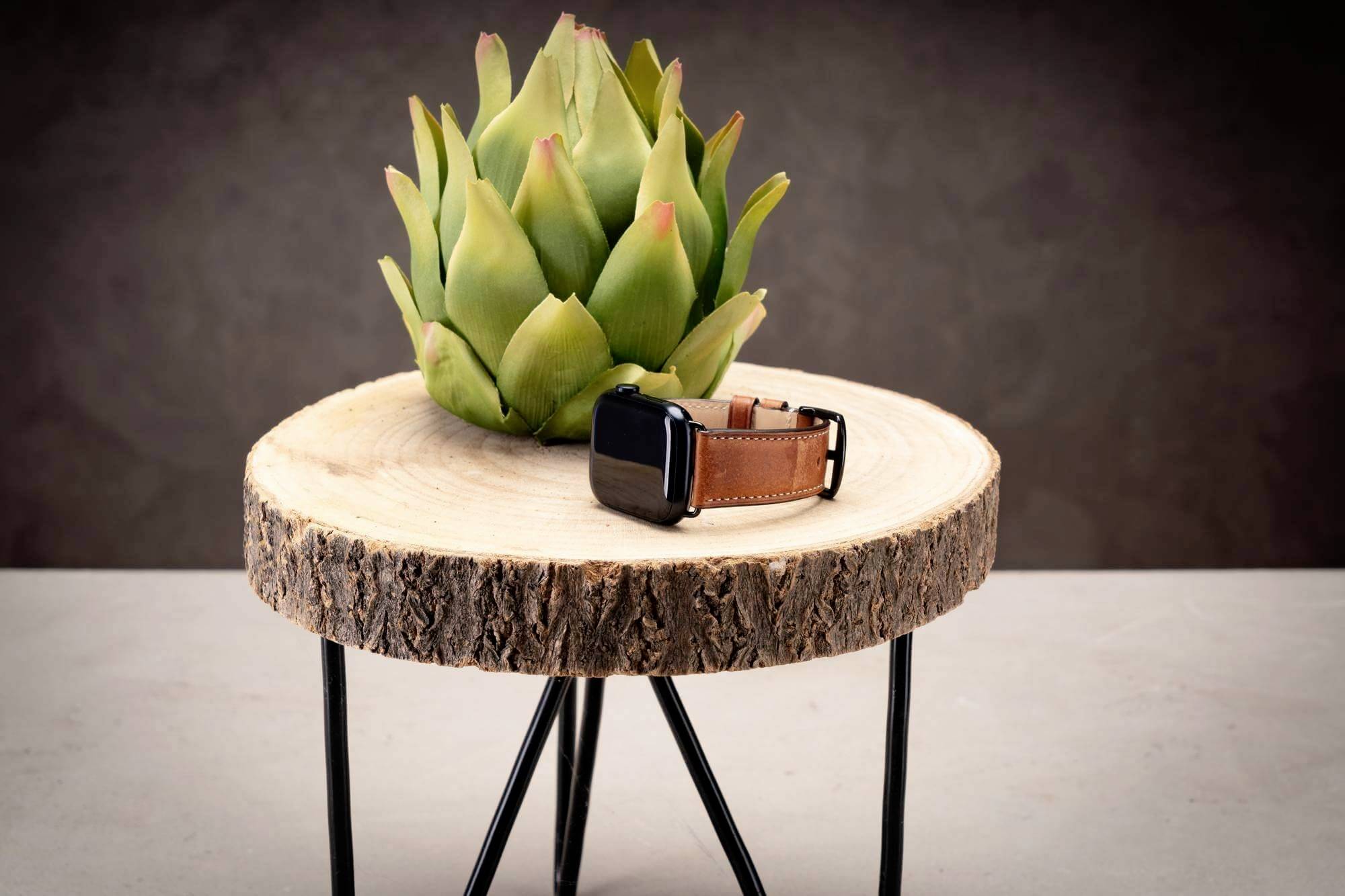 Ein grüner Artischocke und eine elegante Armbanduhr mit Lederband auf einer runden Holzscheibe, platziert auf einem dreibeinigen Tisch.