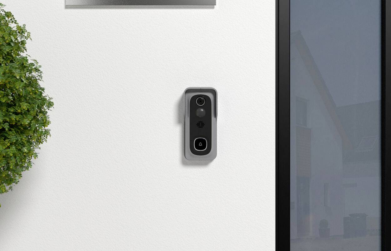 Eine smarte Türklingel mit Kamera ist an einer weißen Hauswand montiert. Rechts ist ein Teil eines Fensters zu sehen, links befindet sich ein Busch.