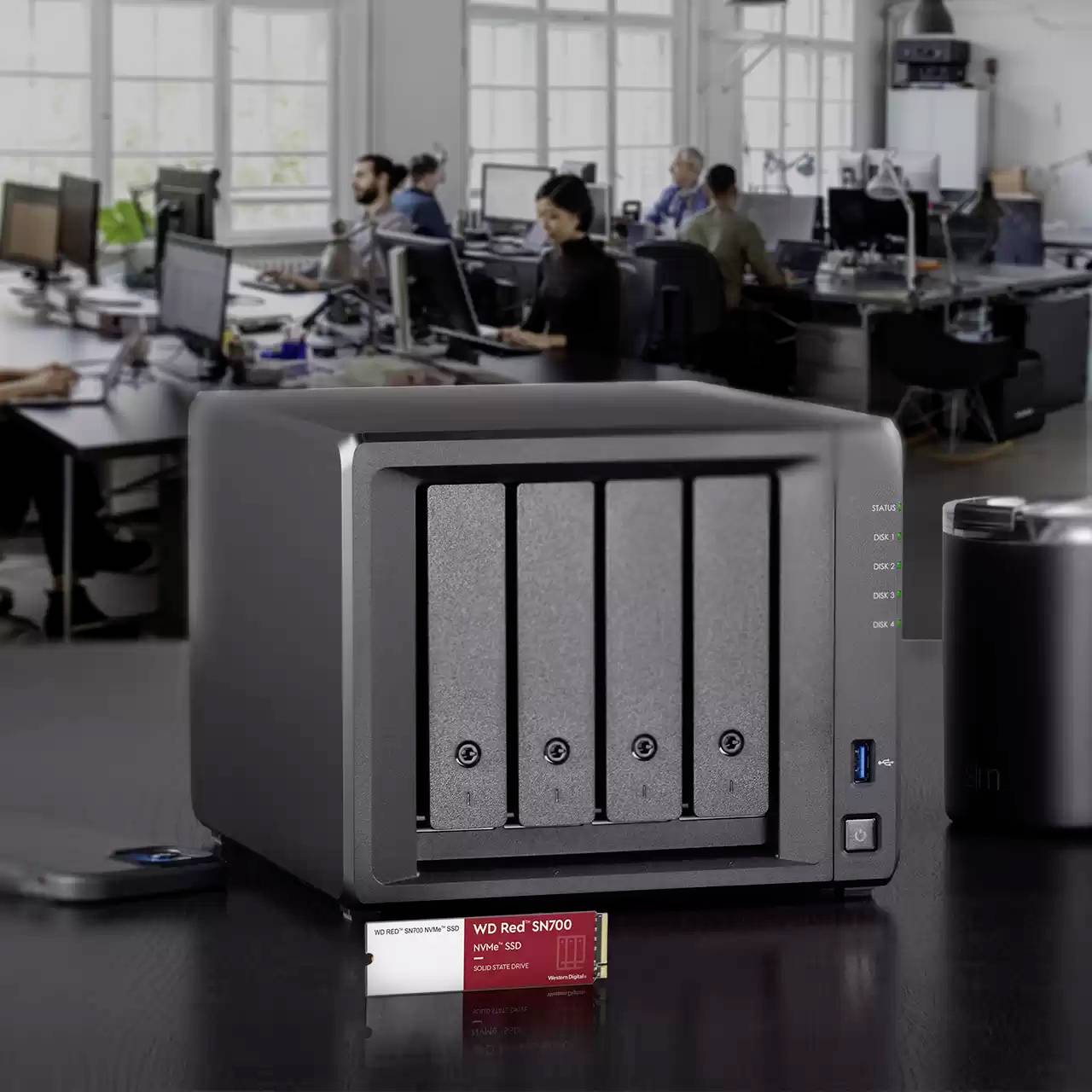 Ein silberner NAS-Server steht auf einem Schreibtisch in einem modernen Büro, umgeben von mehreren Personen an Computern. Im Vordergrund eine WD Red Festplatte.