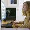Eine Frau sitzt lächelnd an einem Schreibtisch und betrachtet Inhalte auf einem Computerbildschirm. Im Hintergrund ist ein Bilderrahmen sichtbar.