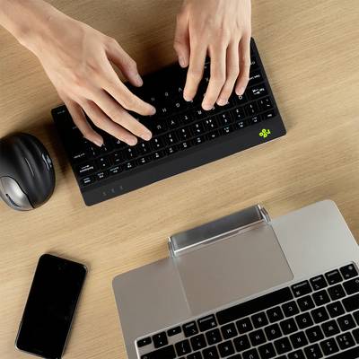 Zwei Hände tippen auf einer schwarzen Tastatur, neben einem Laptop. Eine Computermaus und ein Smartphone liegen auf dem Holztisch.
