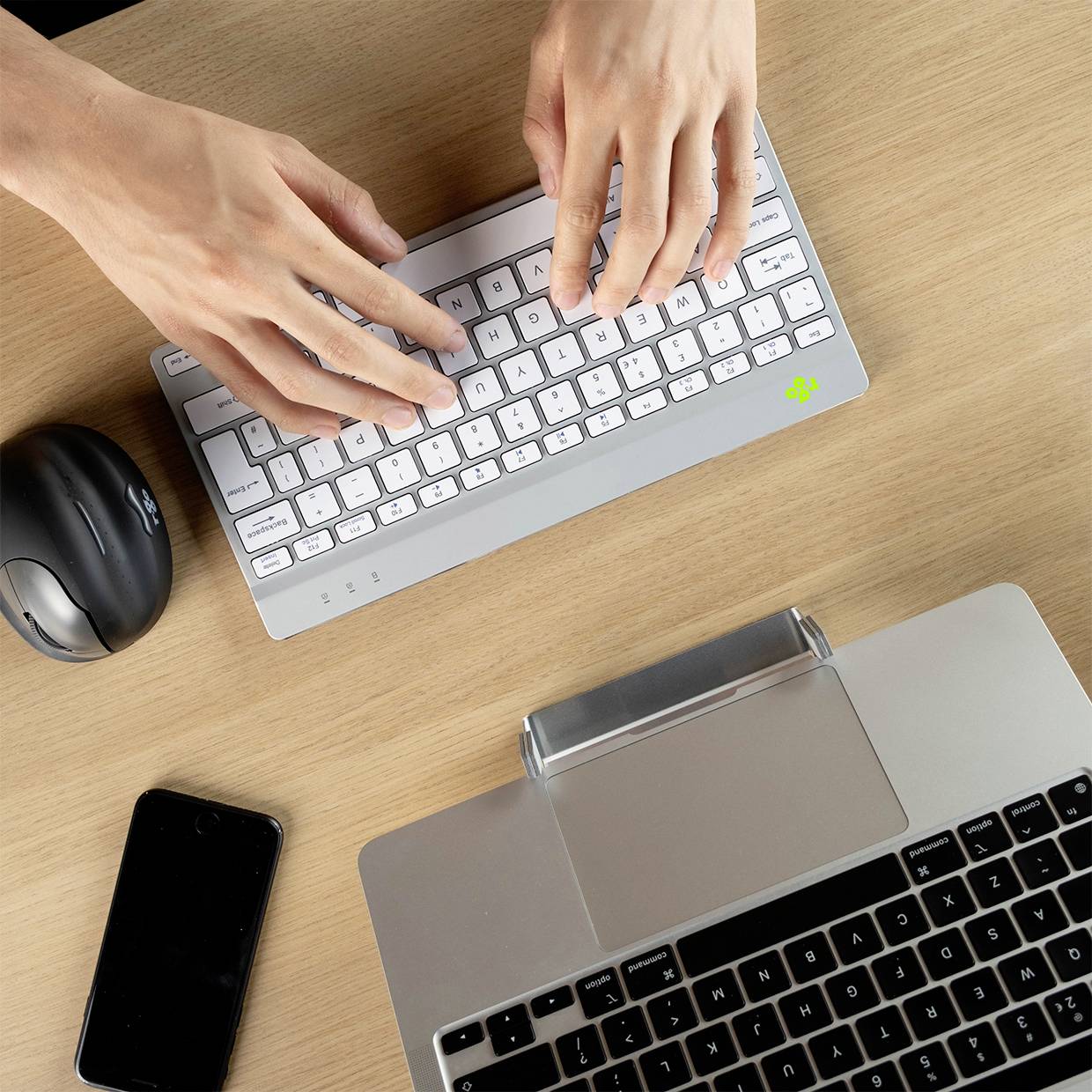 Hände tippen auf eine kabellose Tastatur neben einem Laptop, einer Maus und einem Smartphone auf einem Holztisch.