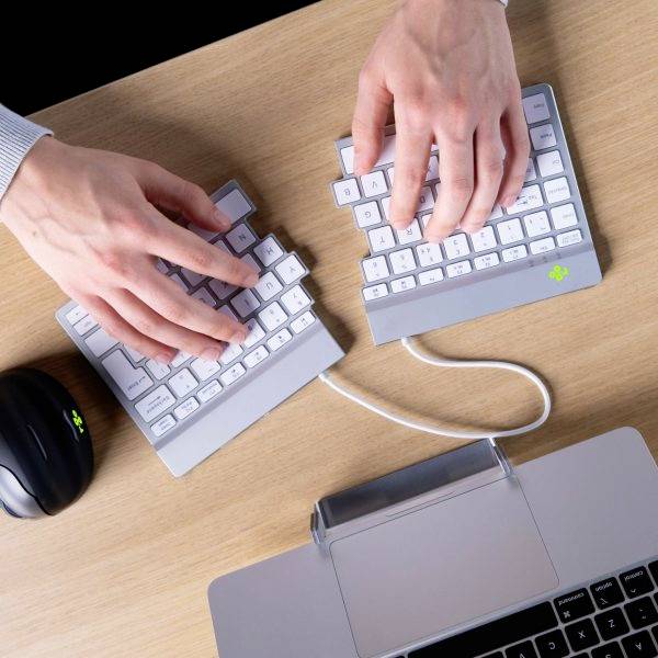 Eine Person tippt auf einer geteilten Tastatur und Maus auf einem Holztisch. Die Hände sind über den Tastaturteilen verteilt.