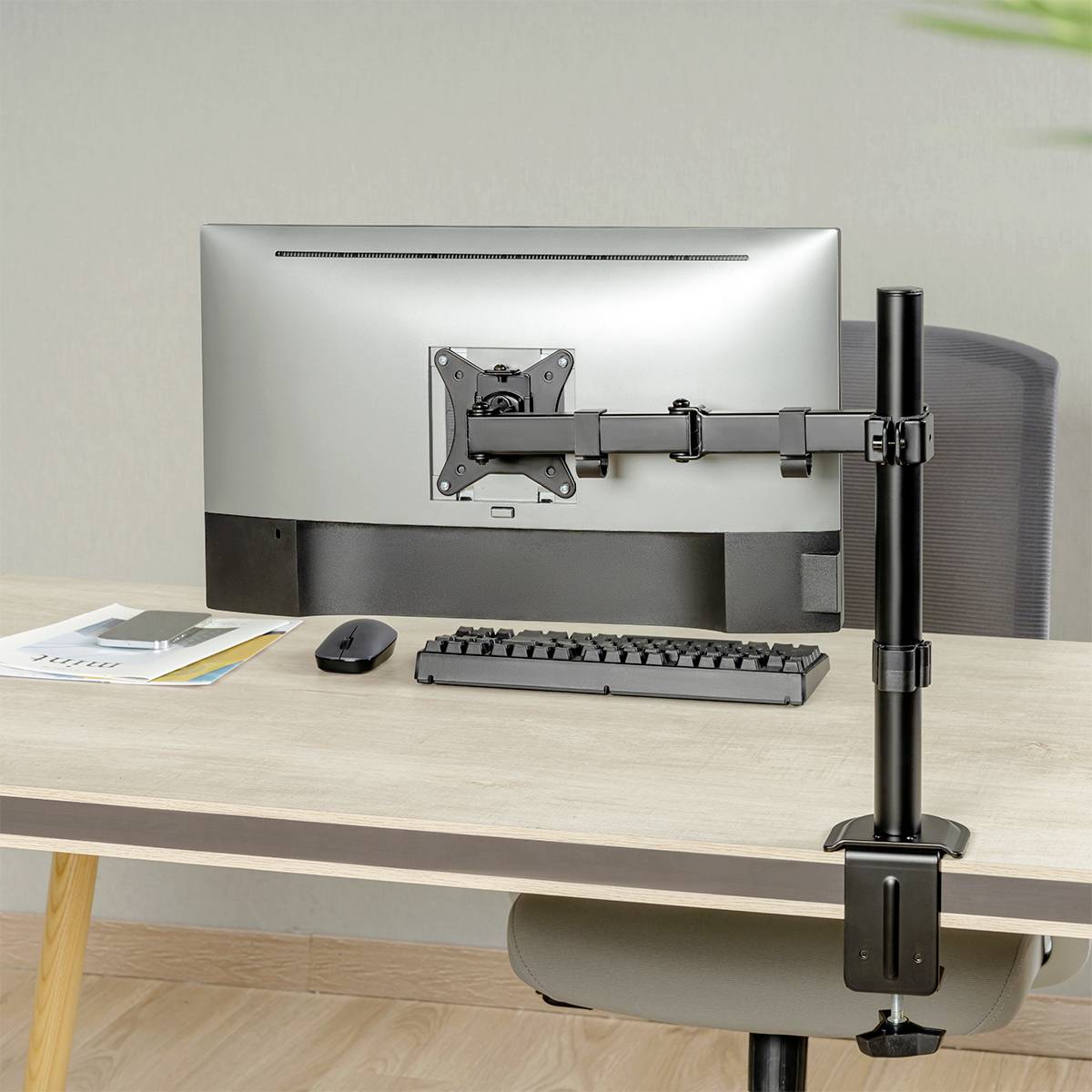 Ein Computerbildschirm ist mit einer verstellbaren Halterung an einem Schreibtisch befestigt. Tastatur und Maus liegen davor.