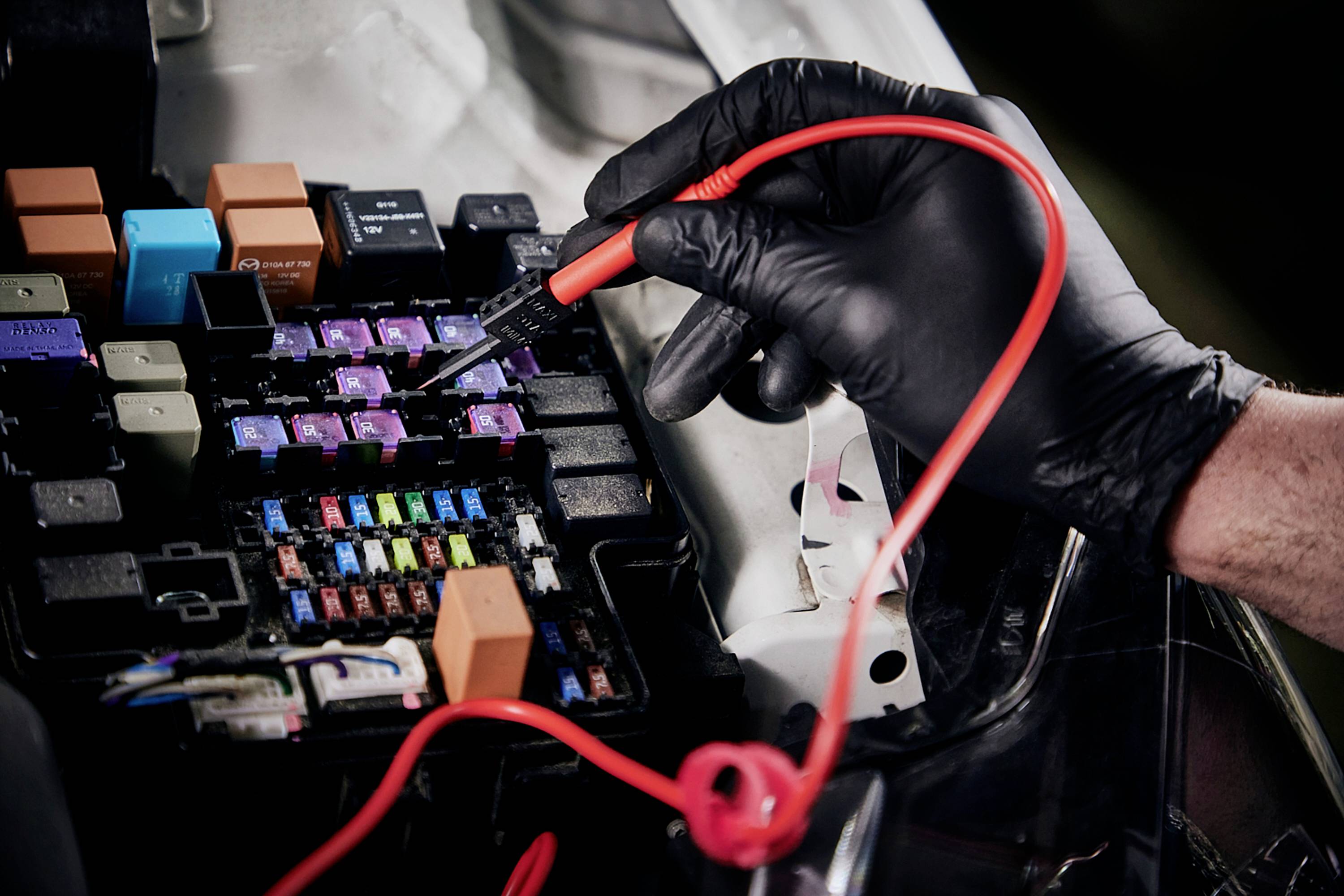 'Ein Mechaniker prüft die Sicherungen in einem Auto mit einem Prüfgerät. Der Fokus liegt auf den farbigen Sicherungen und dem Prüfwerkzeug.'