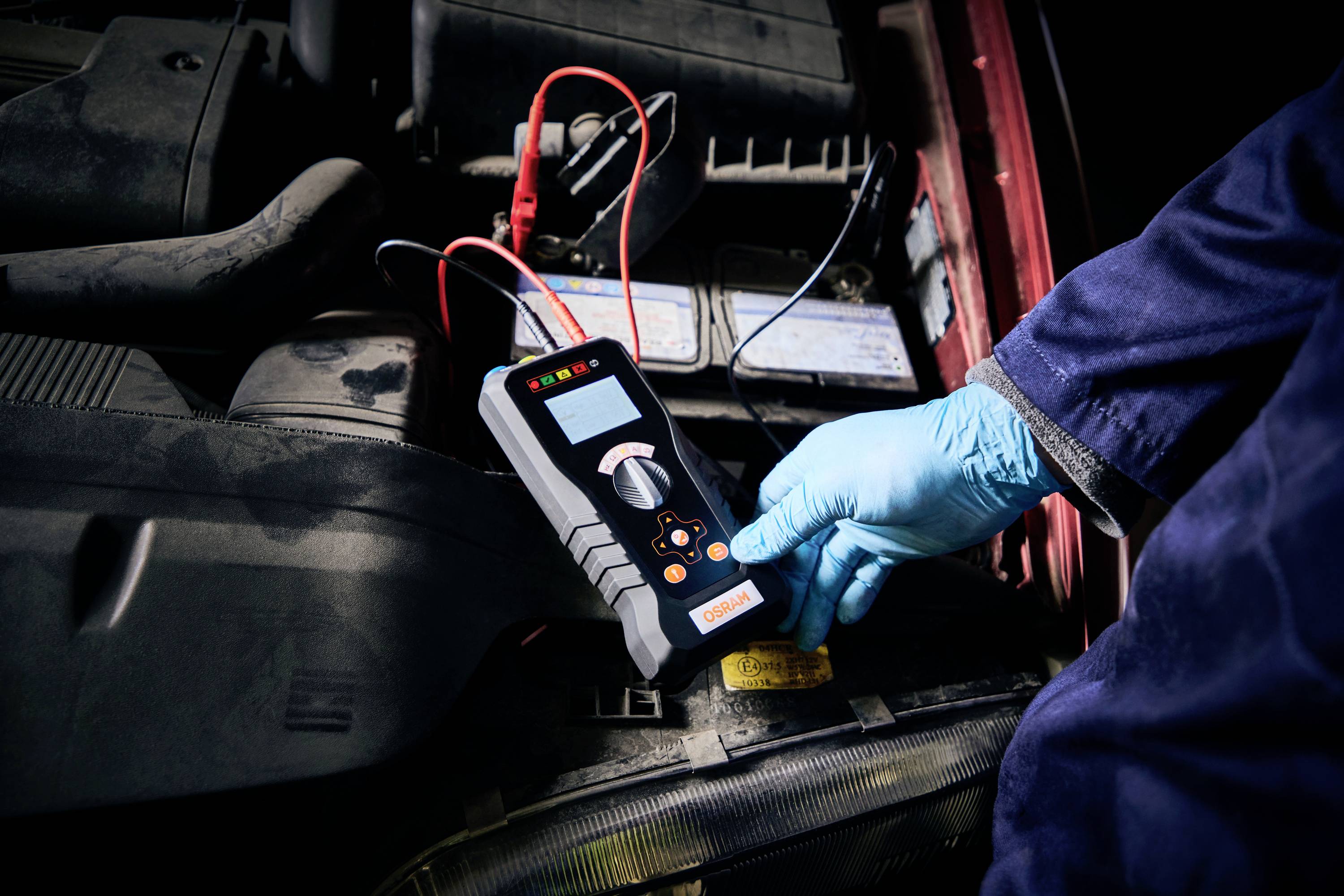 Mechaniker mit blauen Handschuhen misst mit einem Diagnosegerät die Spannung einer Autobatterie im Motorraum.