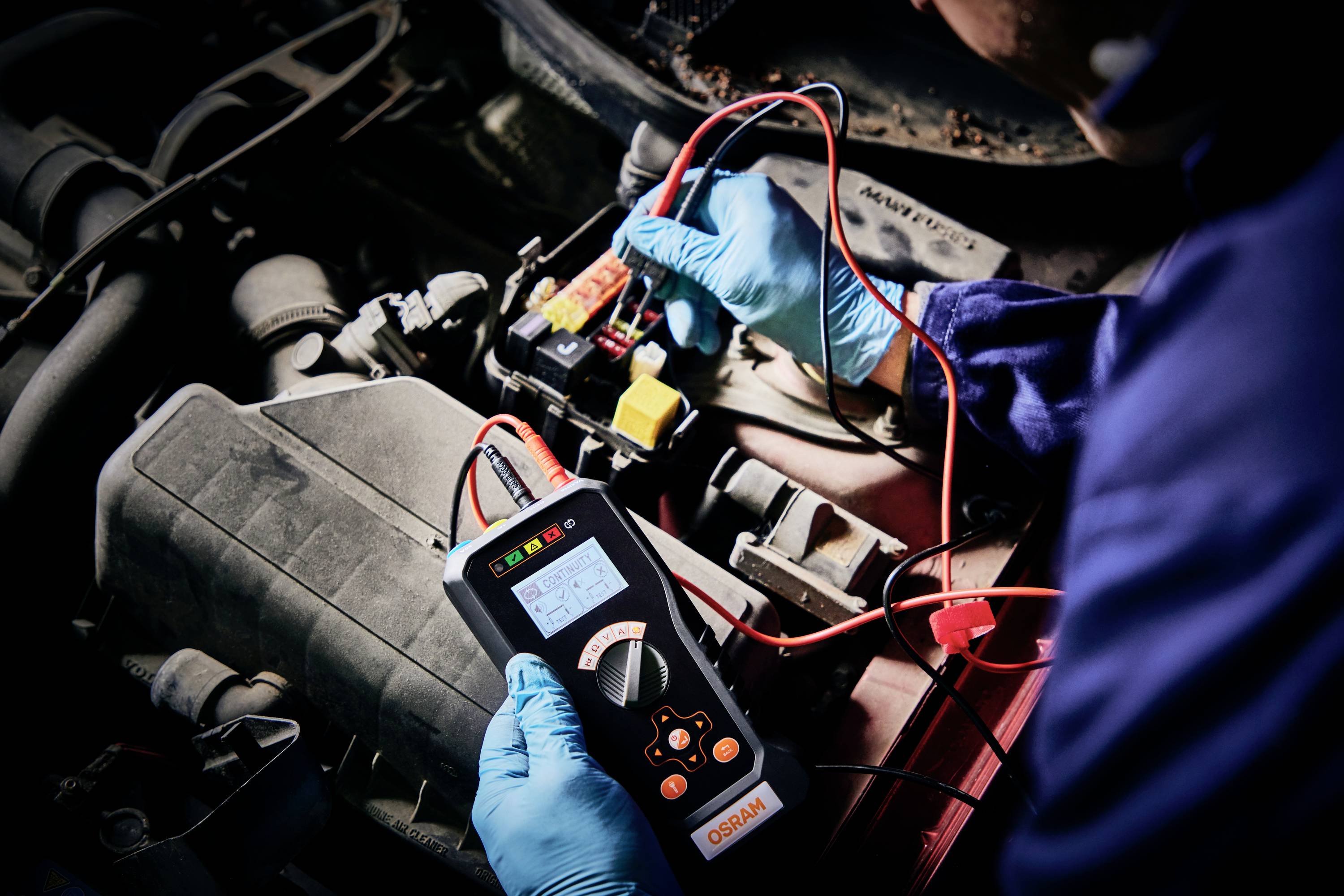 Mechaniker testet Autobatterie mit einem elektronischen Prüfgerät. Hände in blauen Handschuhen halten die Prüfleitungen am Autobatterieanschluss.