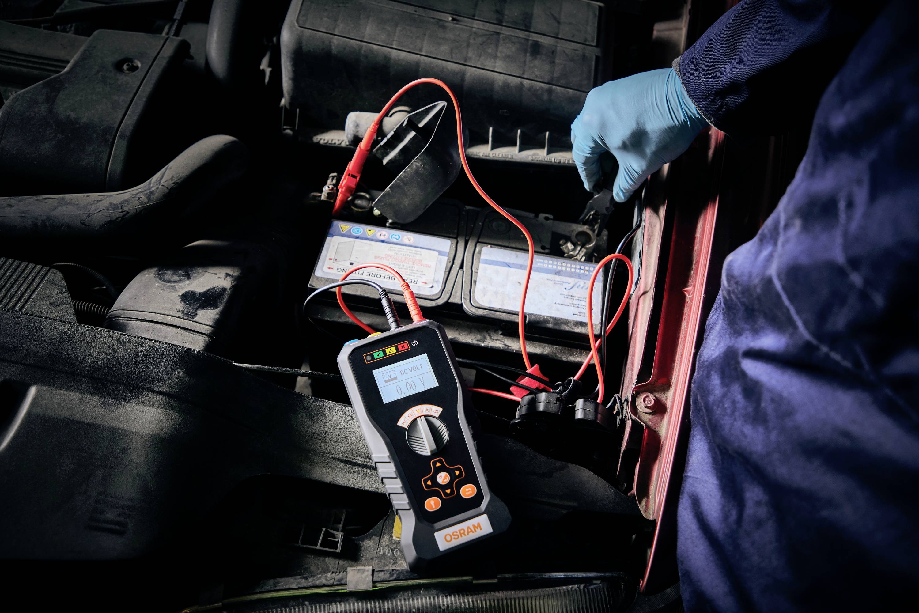 Eine Person mit blauen Handschuhen testet eine Autobatterie mit einem digitalen Diagnosetool. Rote Kabel sind angeschlossen.