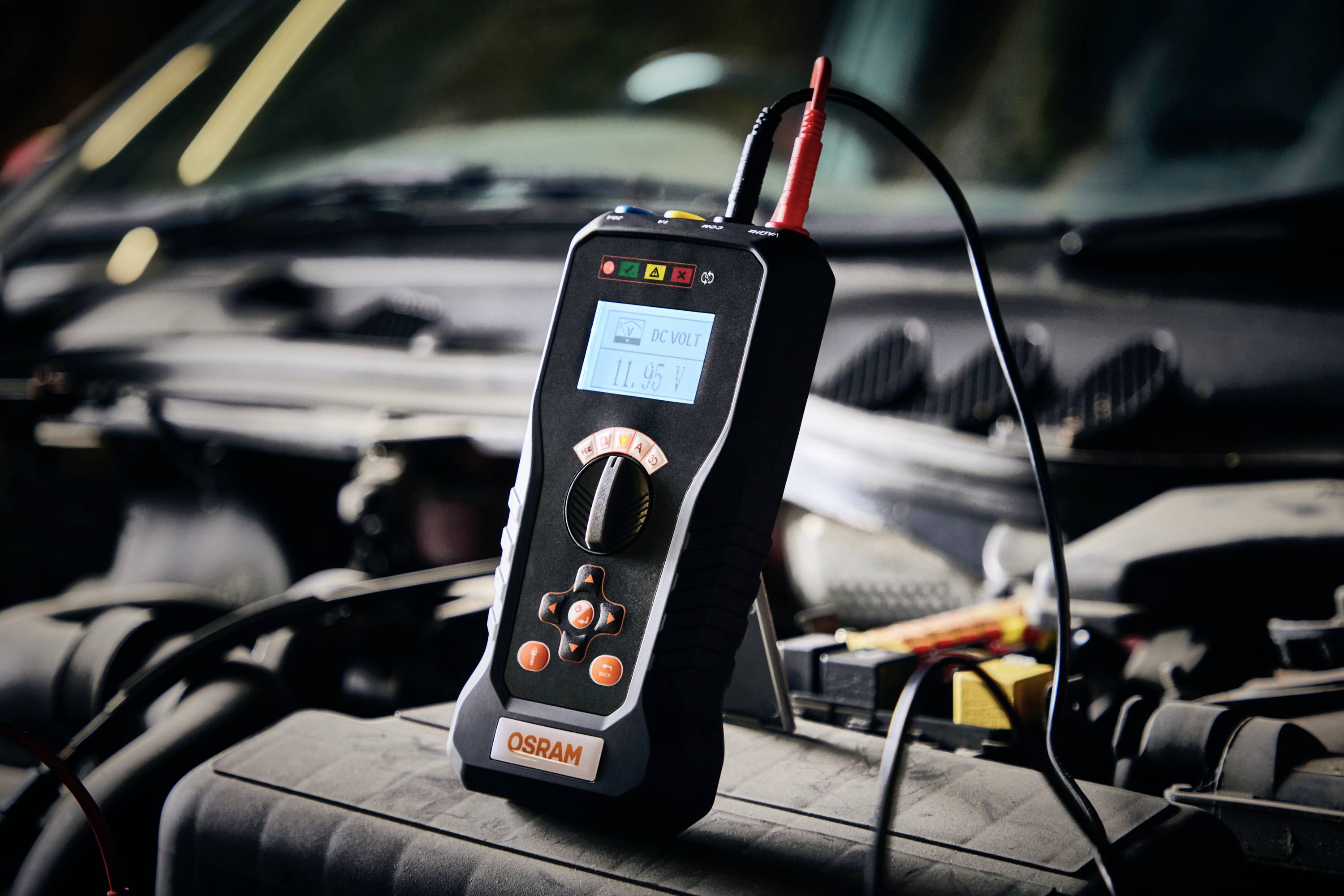 Ein digitales Multimeter misst die Spannung einer Autobatterie unter der geöffneten Motorhaube. Das Display zeigt '11.96V' an.