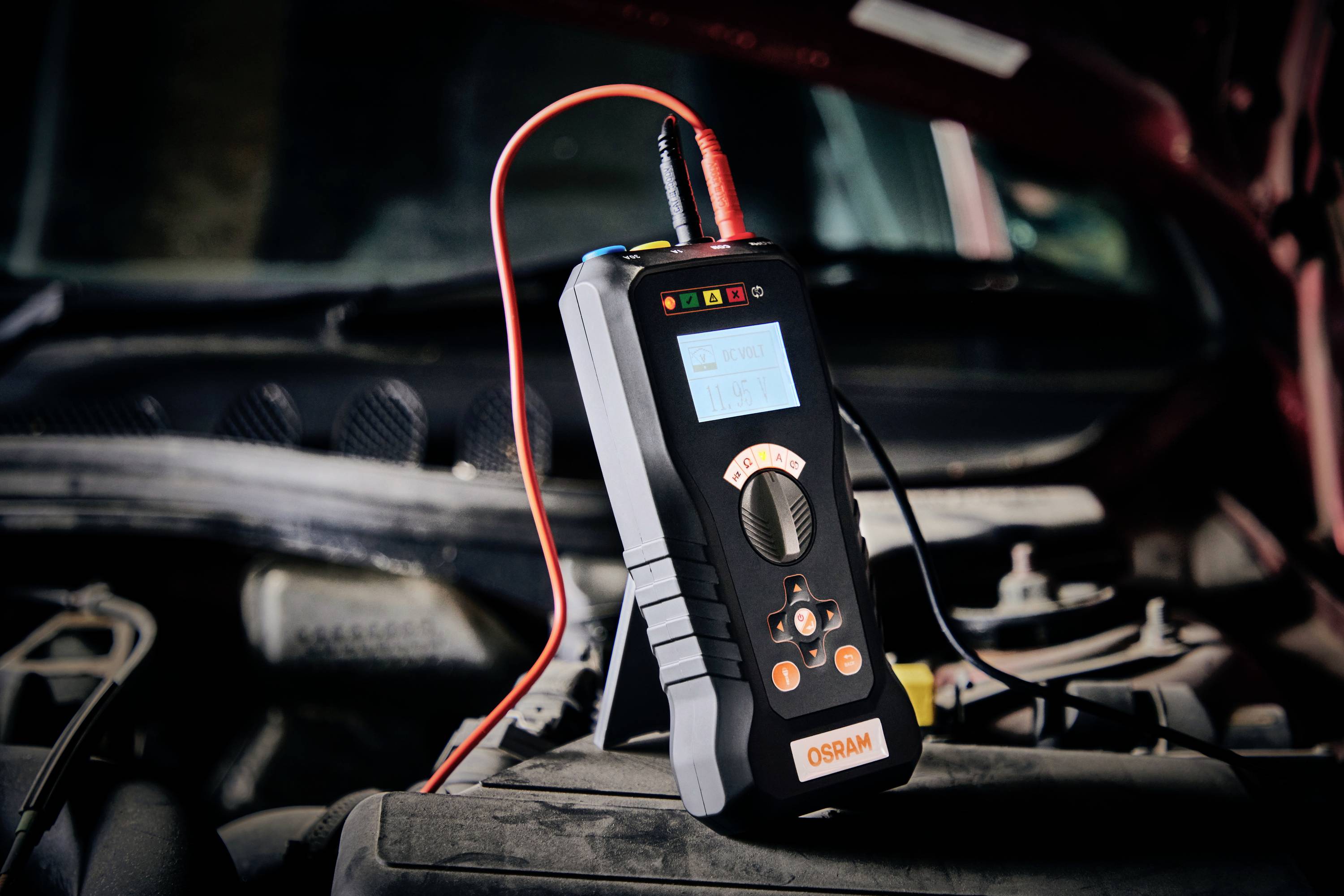 Ein Diagnosegerät mit Kabeln ist an den Motorraum eines Autos angeschlossen. Es zeigt verschiedene Statusleuchten und ein Display.