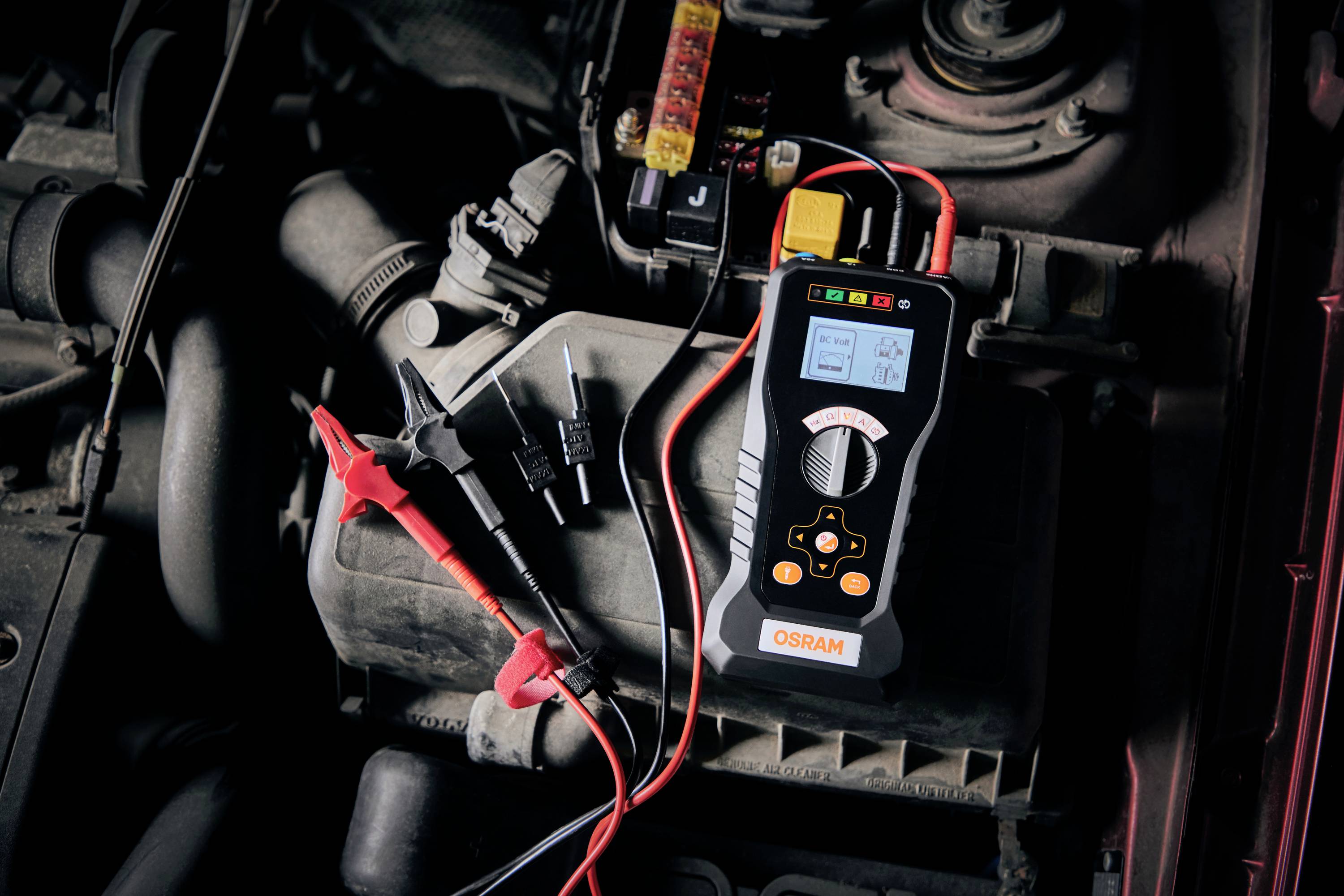 Ein Diagnosegerät zeigt Messergebnisse an und liegt auf einem Auto-Motorblock. Es hat rote und schwarze Kabel mit Klemmen.