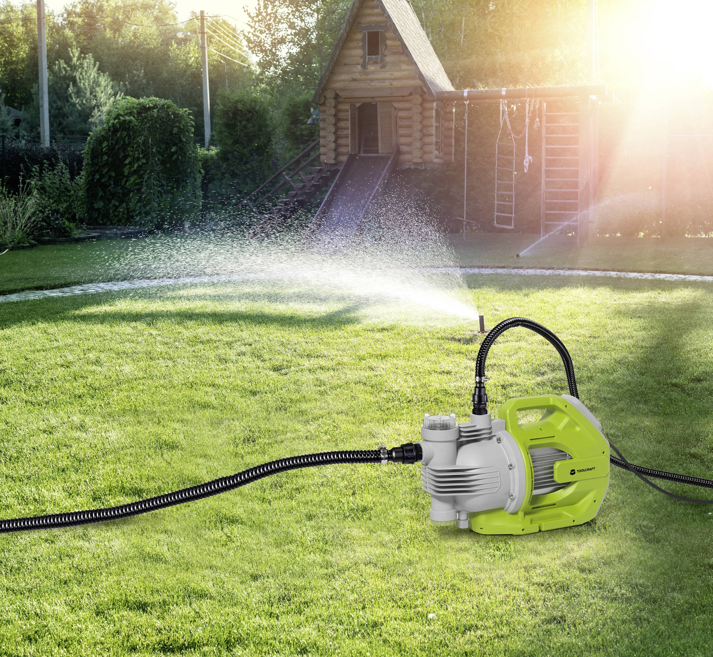 Grüne elektrische Wasserpumpe auf einem Rasen in einem Garten, bewässert den Rasen mit einem feinen Wasserstrahl; Holzhütte im Hintergrund.