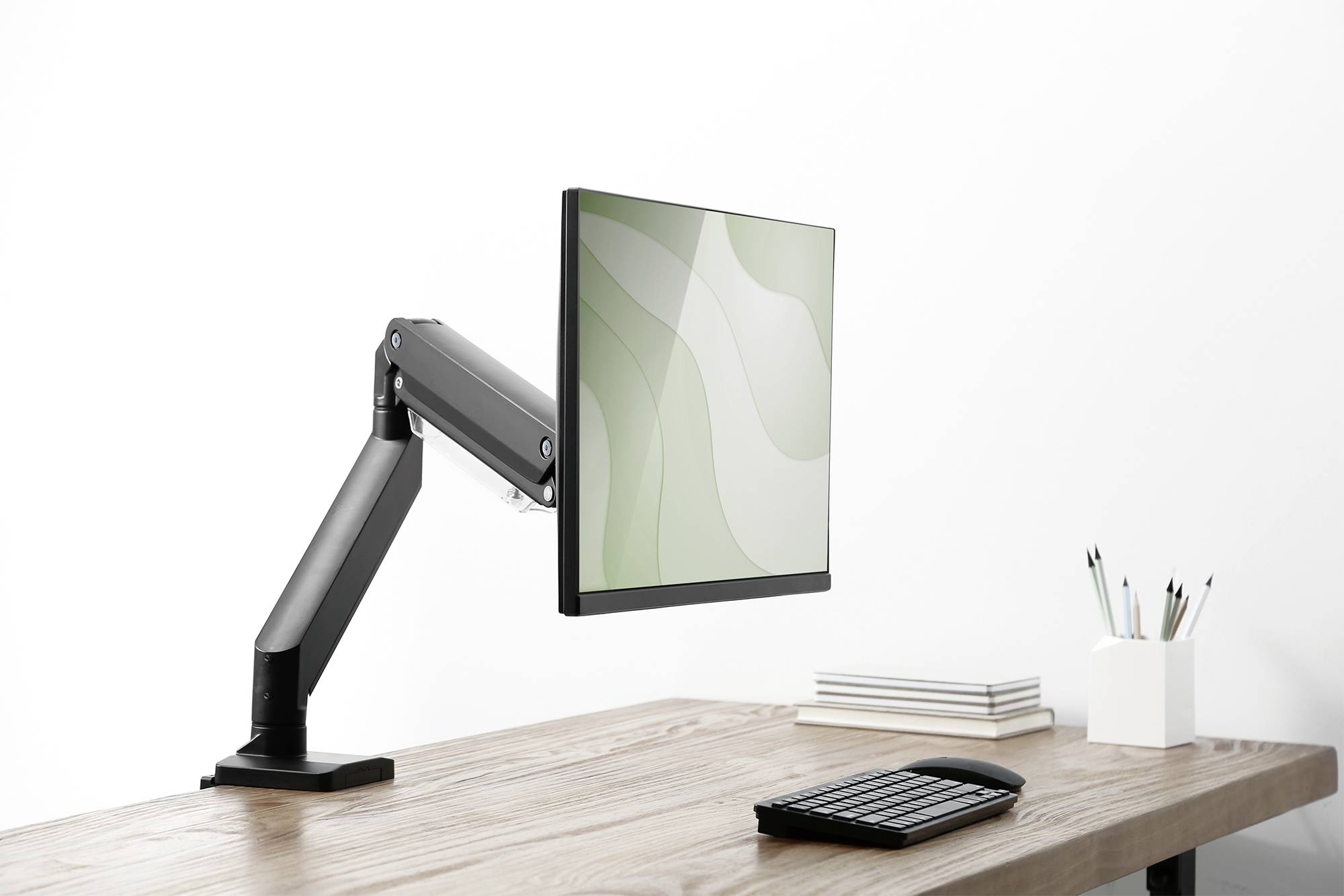 Ein Monitor an einem verstellbaren Arm auf einem Schreibtisch mit Tastatur und Stiftebecher. Im Hintergrund neutrale Wand.
